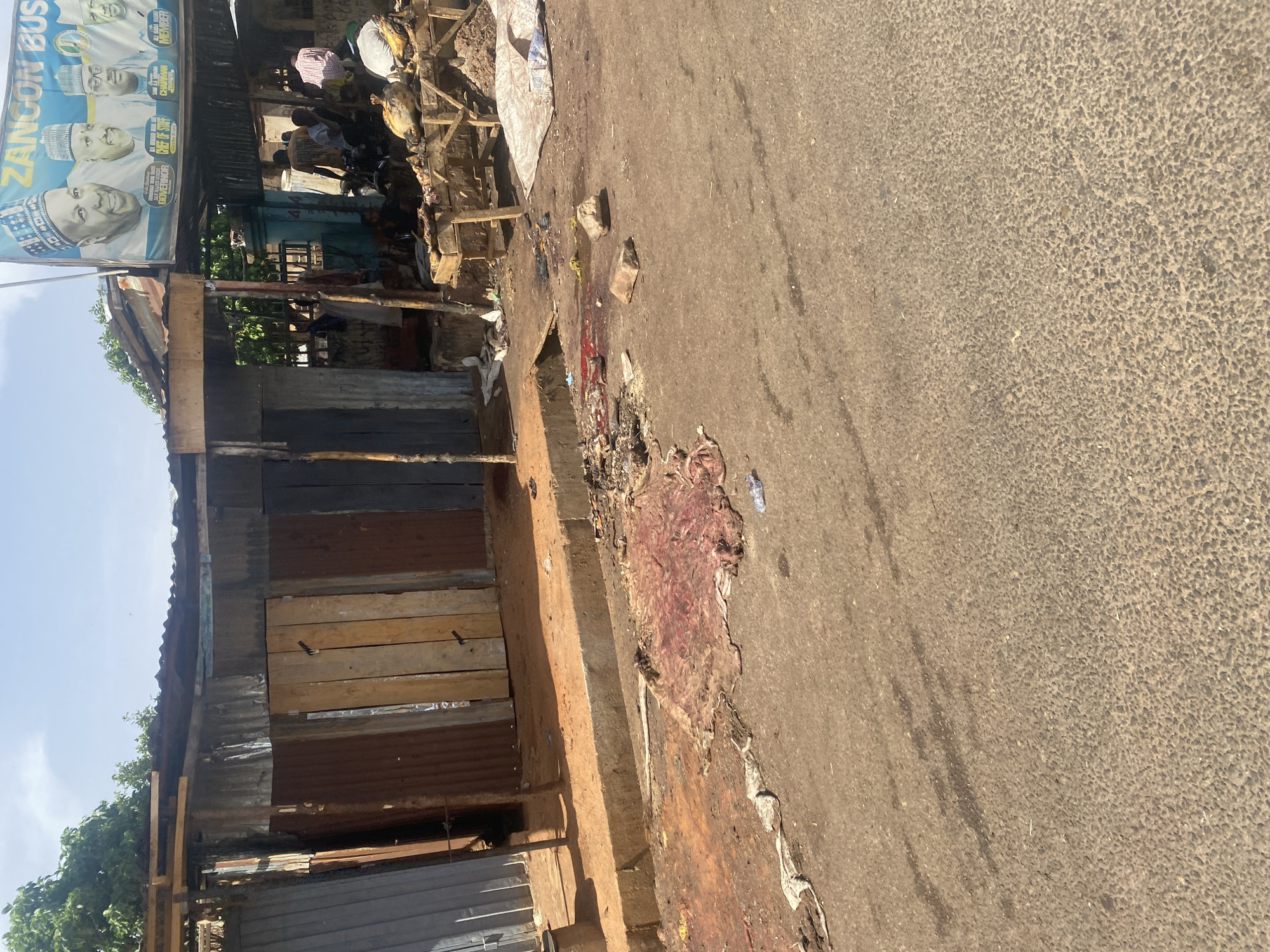 A street scene in Gombe, Nigeria, depicts a building with corrugated metal and wooden plank walls. A political campaign banner featuring the images of multiple men is attached to the building's facade. Adjacent to the building, the paved street displays stains and discarded material, including a large, reddish substance resembling animal hide or entrails. Several individuals are visible under a canopy structure to the left of the street. The road surface is rough and light-colored.