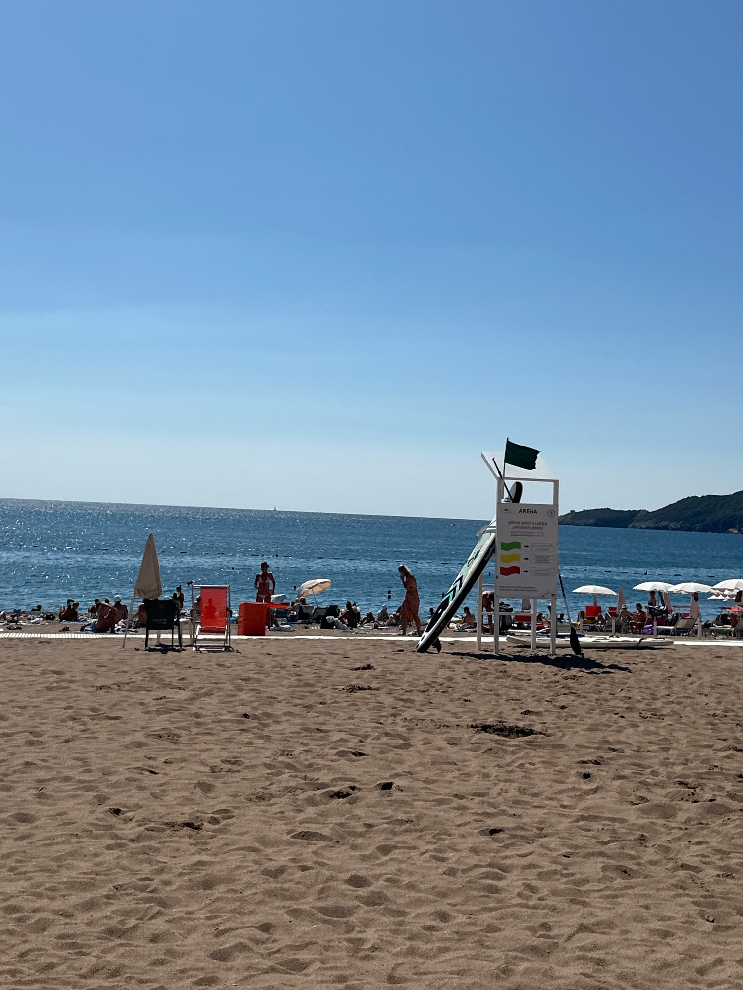 The image depicts a sandy beach in Budva, Montenegro, on a clear, sunny day. The foreground is dominated by the textured sand of the beach, with footprints and subtle undulations. In the midground, the beach is populated with numerous individuals relaxing on the sand or near the water. Beach chairs, umbrellas, and towels are scattered across the sand. A prominent lifeguard tower with a green flag and informational signs stands near the water's edge. The water, a deep blue, stretches to the horizon, with gentle waves lapping the shore. In the background, a lush, green hillside slopes down to the sea, with buildings visible at its base. The sky is a vibrant, cloudless blue, indicative of fair weather.
