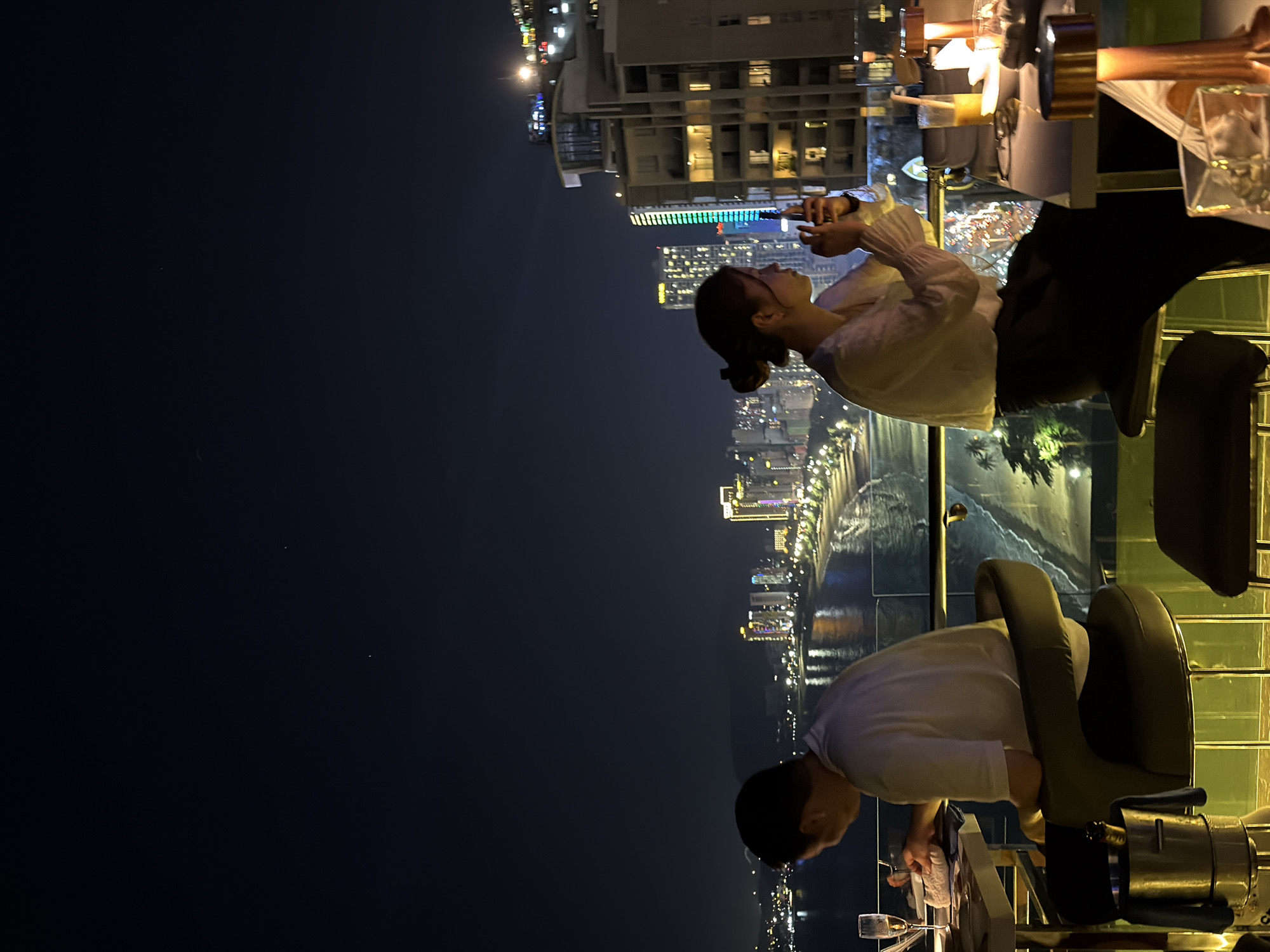 Here's a description of the image:
**Scene:** The image is taken at night, likely from a high vantage point in Nha Trang Ward, Vietnam, overlooking the city and coastline. The skyline features lit buildings, including skyscrapers.
**People:** There are two people visible: a woman sitting and using her phone, and a man standing next to a table. The woman has her hair up in a bun.
**Objects:** A table with drinks, glasses, and ice bucket is present. There are chairs at the table. A clear railing protects the edge of the location.
**Actions/Interactions:** The woman is interacting with her phone. The man appears to be tidying up.
**Environment:** The night sky is dark. The cityscape below is illuminated with lights, and there are signs of traffic along the coastline. The lighting in the immediate area is artificial.