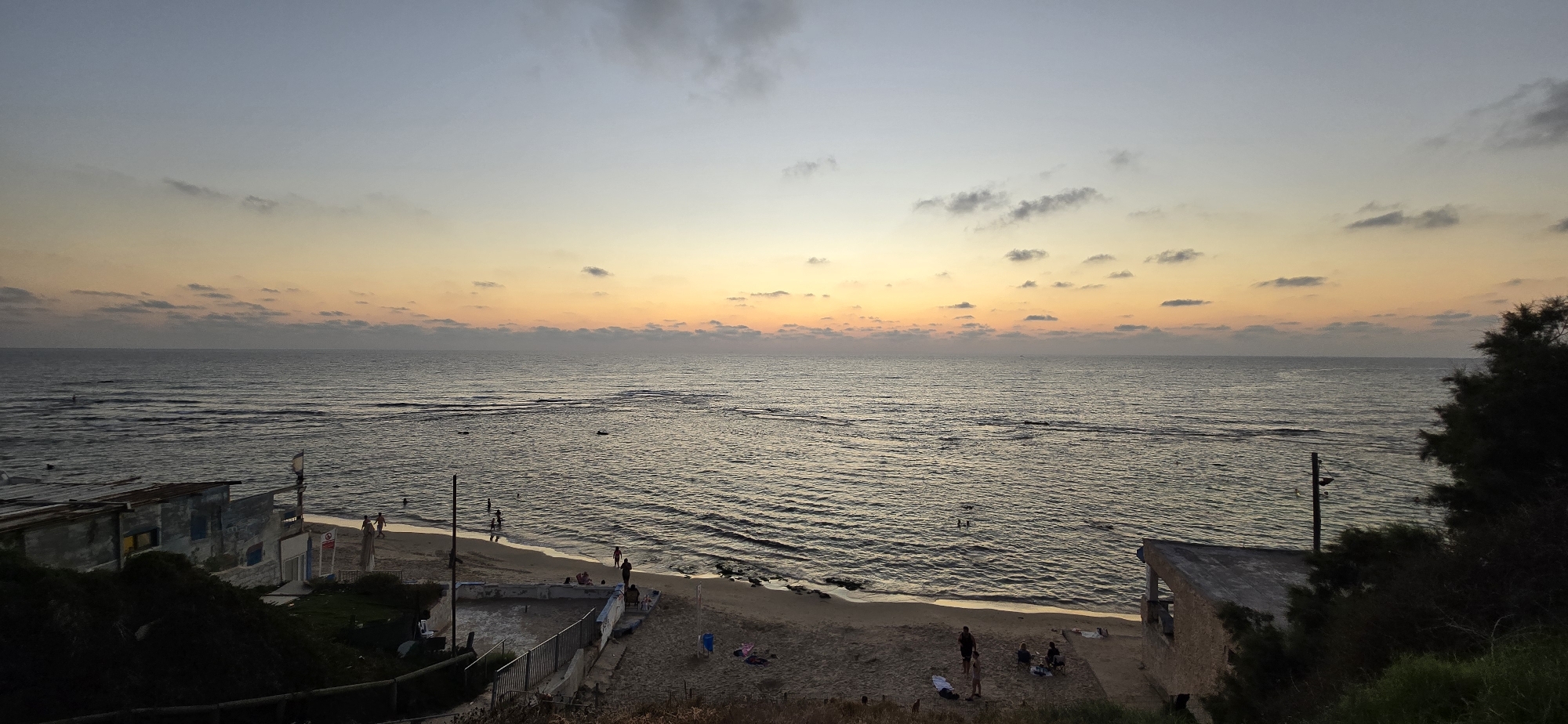 This is a wide-angle, eye-level photograph taken from a slightly elevated viewpoint overlooking a beach scene at sunset. The sky displays a gradient of colors, transitioning from a pale blue at the top to warm oranges and yellows near the horizon, with scattered clouds adding texture. The calm sea reflects the sky's hues, with gentle waves lapping at the shore.
On the sandy beach, several people are visible, engaged in various activities. Some are wading or swimming in the shallow water, while others are relaxing on the sand. There are a few small, simple structures along the beachfront, including what appears to be a makeshift changing room or small building with a corrugated metal roof on the left, and a more substantial, partially constructed building with an open roof on the right. Beach furniture, like chairs and towels, are scattered on the sand, suggesting a leisurely atmosphere. The overall mood is tranquil and serene, indicative of the end of a day at the beach.
The time of day is clearly sunset, given the sky colors and the soft lighting. The weather appears calm and pleasant, suitable for beach activities. The presence of the structures and the specific type of vegetation (a dark, bushy tree on the far right) might offer subtle clues to the location. The scene evokes a sense of peace and relaxation. No visible text is discernible in the image.