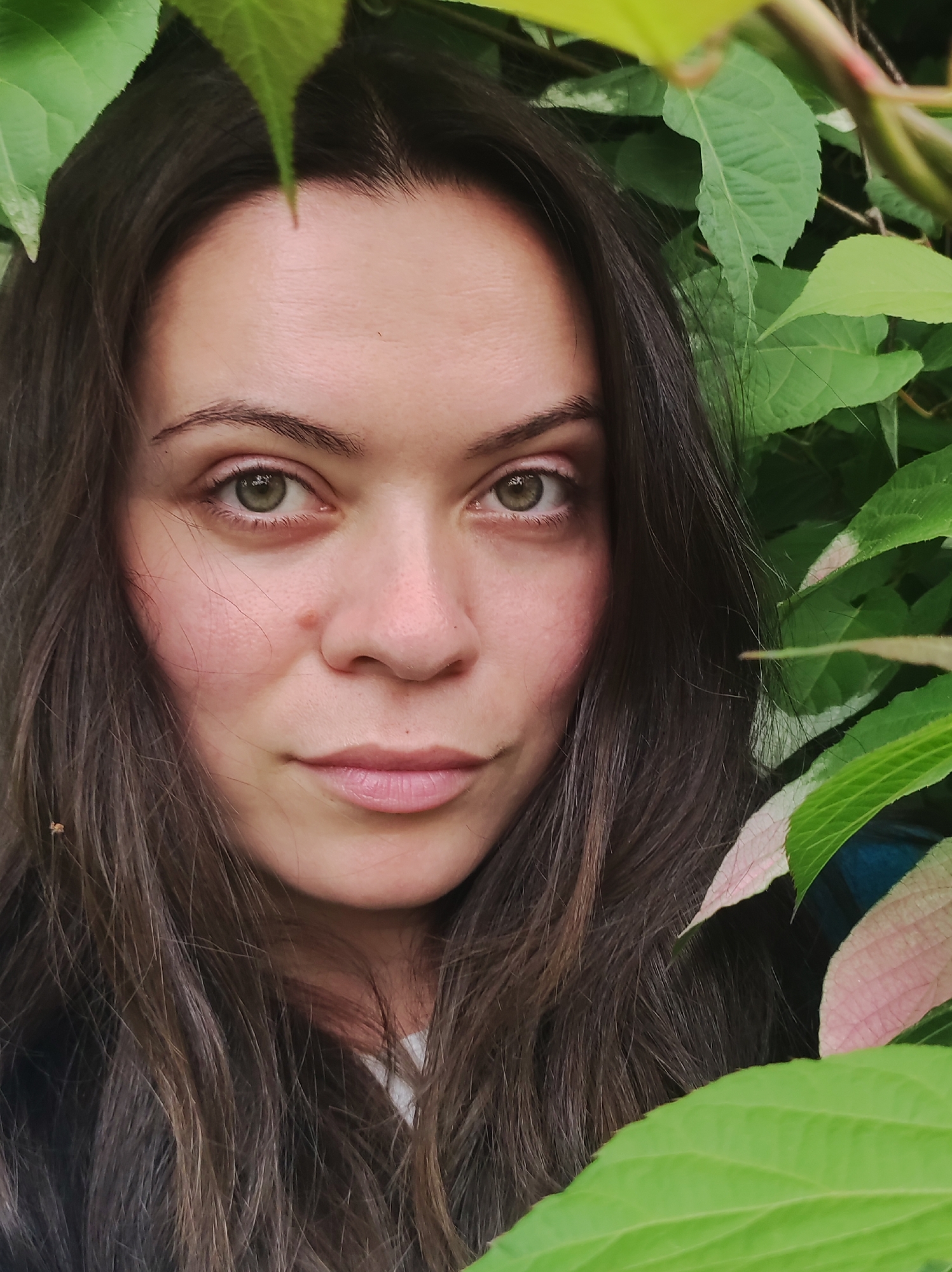 A close-up, eye-level shot features a woman with long, dark brown hair partially obscured by lush green foliage. She is positioned centrally in the frame, her face angled slightly towards the viewer. Her eyes are a striking green, and her expression is neutral. Her skin appears fair with a rosy blush on her cheeks. The background is dominated by vibrant green leaves, some with pinkish hues, suggesting an outdoor, natural setting. The image does not provide any specific visual cues to indicate a location within the city of Fokino, Russia.