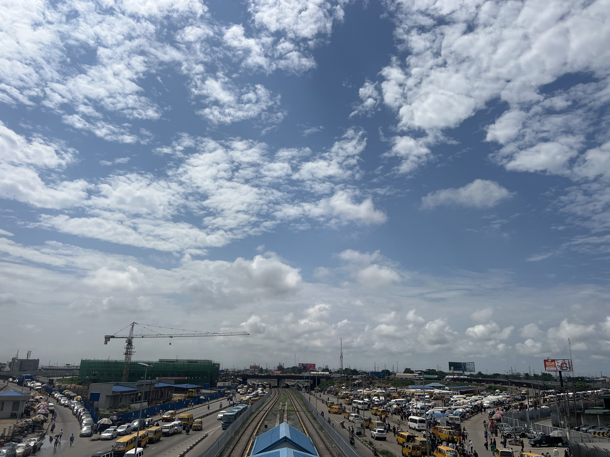 The image captures a bustling urban scene under a partly cloudy blue sky. In the foreground, a wide road is filled with an array of vehicles including numerous yellow minibuses, white vans, and a few cars. Pedestrians navigate the sidewalks and the edges of the road, some under colorful umbrellas. To the left, a line of parked vehicles is visible, with market stalls and awnings set up.
Further into the scene, train tracks run parallel to the road, with a blue-roofed station structure situated between them. A long train is stopped on the tracks. In the middle ground, a large construction site is evident, with a prominent crane towering over buildings draped in green netting. Beyond the construction, the cityscape stretches out, with various structures, billboards, and utility poles visible against the horizon. The environment appears to be a busy transportation hub and commercial area.