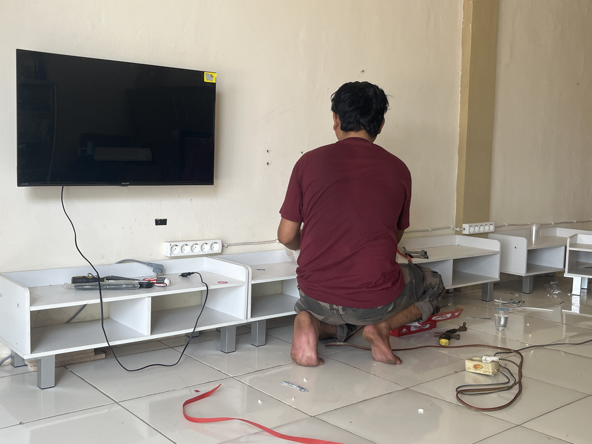 A man, viewed from behind, is kneeling on a tiled floor in a room. He is wearing a maroon t-shirt and camouflage-patterned shorts. He is working near a white media console with multiple shelves, which is positioned against a cream-colored wall. A black flat-screen television is mounted on the wall above the console. Several electrical cords and power strips are visible on the floor and the console. Tools, including pliers and a hammer, are also present. Red tape is visible on the floor. The environment appears to be an indoor space in Sekayu, Indonesia, likely a room undergoing installation or setup of the media equipment.