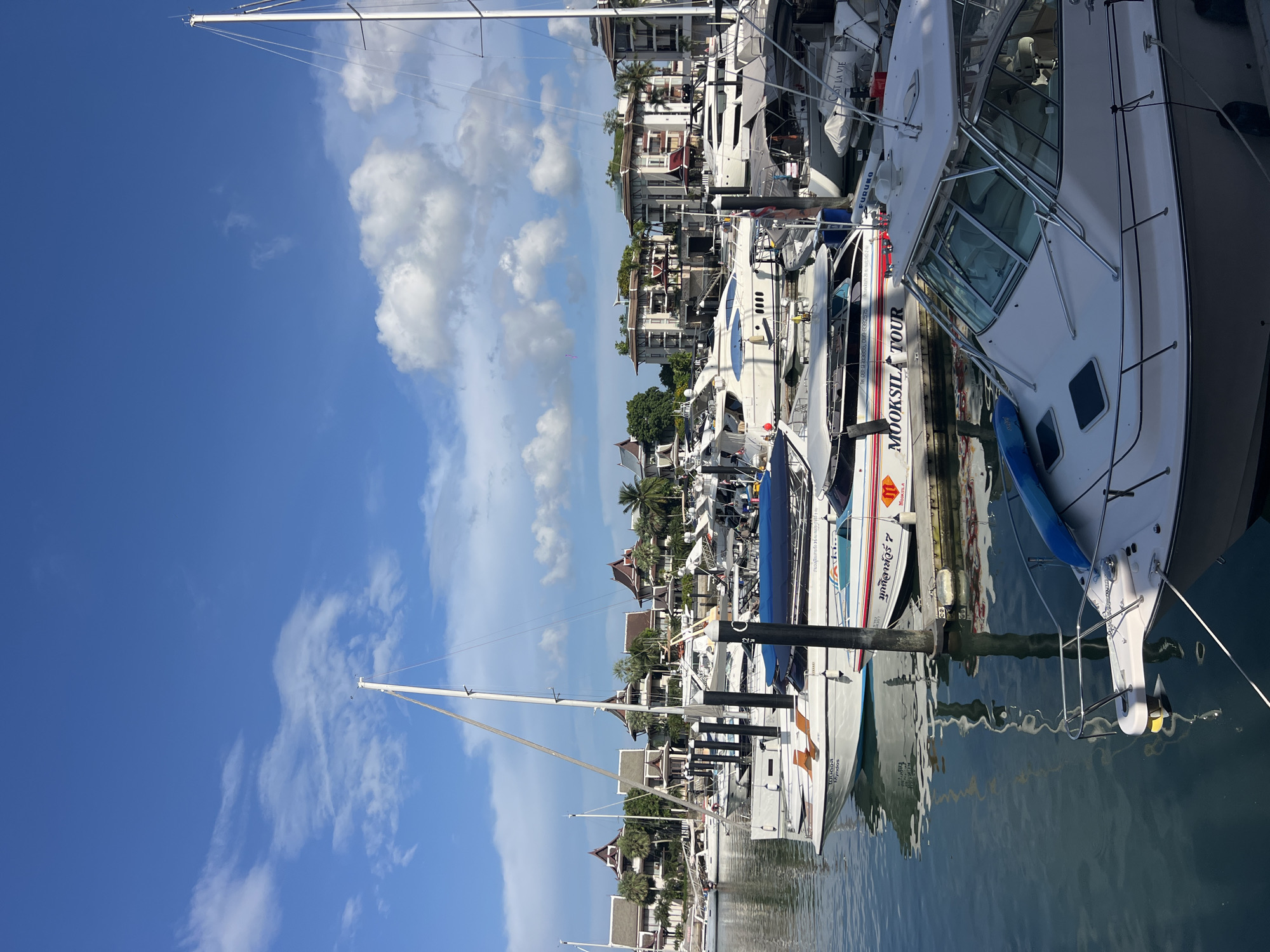 The image shows a marina with several boats docked in calm water. The boats are of various sizes and types, including sailboats and motor yachts. In the background, there are buildings with red-tiled roofs, palm trees, and a blue sky with white clouds. The marina appears to be in a tropical or subtropical location, possibly a resort area. The scene is serene and picturesque, with the sunlight reflecting off the water and the boats. The location is identified as Ko Kaeo, Thailand, a known area with marinas and tourist attractions.