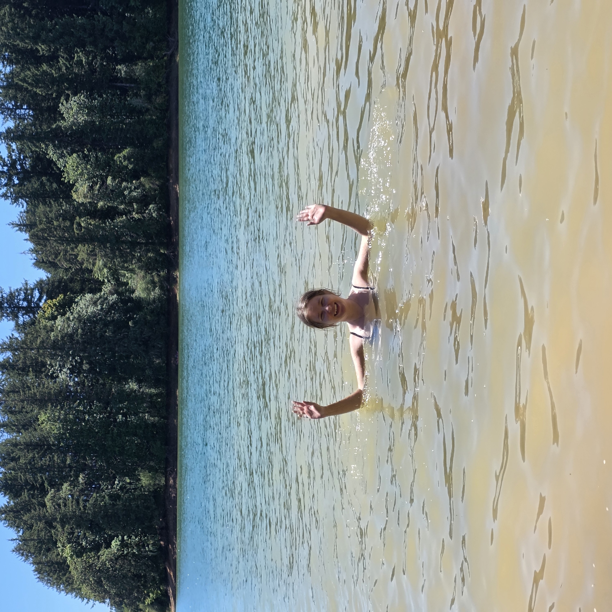 In Cherry Grove, United States, a young woman is swimming in a lake. She is partially submerged in the water, with her upper body visible. Her arms are raised in the air, and she is smiling at the camera. The water is clear and calm, with sunlight reflecting off its surface. In the background, a dense forest lines the edge of the lake. The sky above is a clear blue.