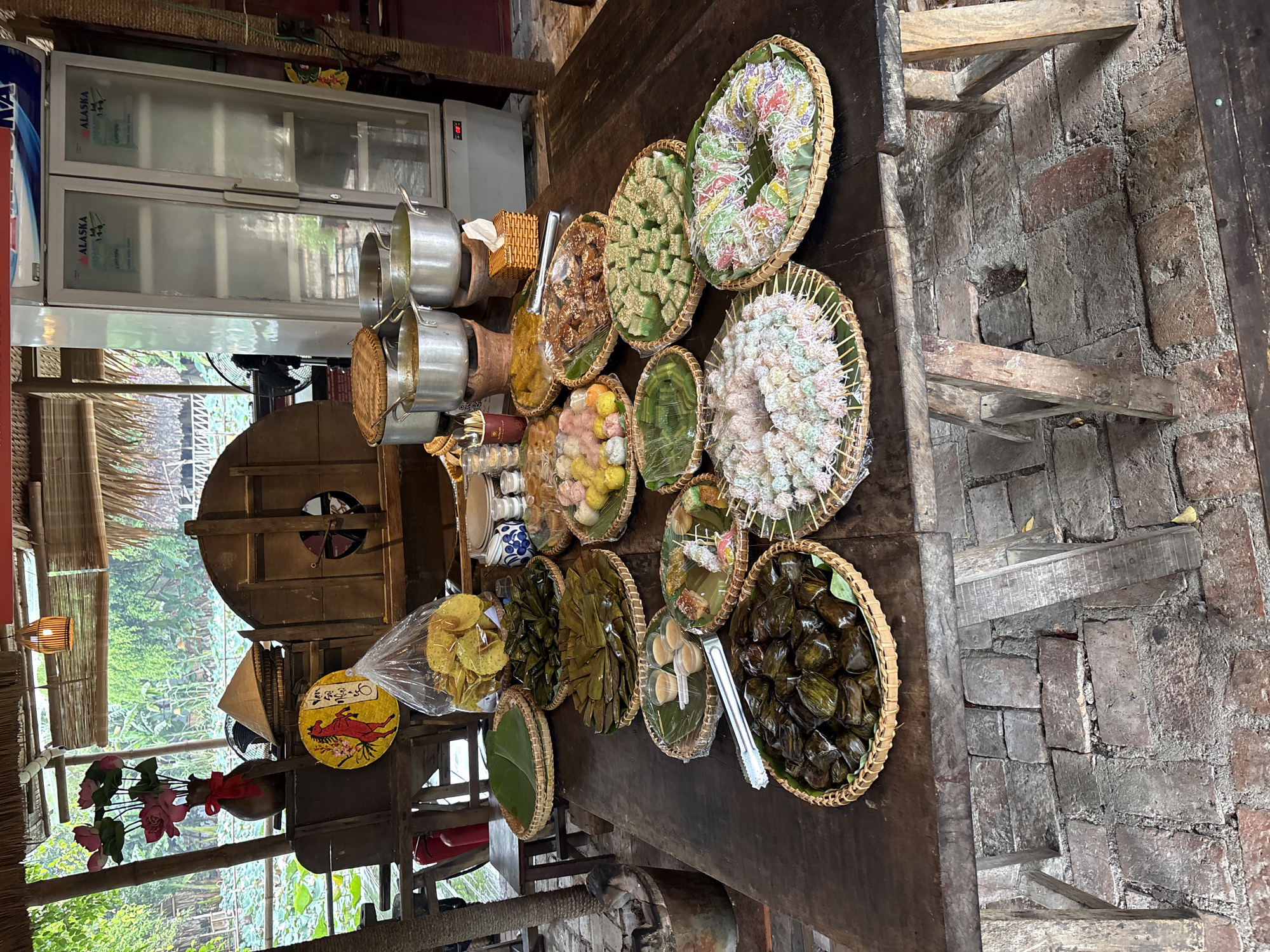 The image captures a variety of traditional Vietnamese desserts and snacks displayed in woven baskets on a long, dark wooden table. This scene is likely located within a shop or market in Nam Nha Trang Ward, Vietnam, as evidenced by the style of the food, decor, and surrounding environment.
Several baskets hold colorful, bite-sized desserts, including what appear to be cakes, candies, and other sweets. The items are arranged neatly and appear to be fresh. In the background, there are cooking implements, suggesting that the location is where the items are prepared or served. Additionally, there is a refrigerated display case.
The environment includes a weathered brick floor and rustic wooden beams, pointing to a traditional style of architecture. There is a sign on the wall with Vietnamese script. The scene suggests a vibrant and local culinary experience in Vietnam.