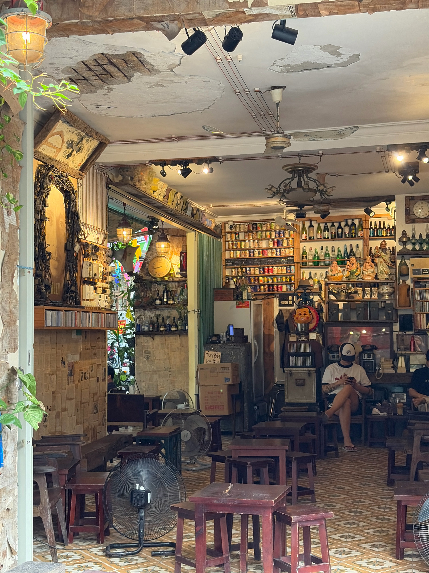 This image depicts the interior of a cafe in Da Nang, Vietnam. The cafe has a rustic and eclectic decor. The walls are covered in newsprint, and there are exposed wires and cracked plaster on the ceiling, with industrial-style lighting fixtures. Shelves are filled with bottles, cans, and decorative items, including Buddha figurines. There are multiple tables and wooden stools scattered throughout the space, with some set up for customers.
In the foreground, a person is seated at a table, looking at their phone. Another person is partially visible in the background. Several fans are strategically placed around the room, indicating the climate. The floor is tiled with a patterned design. The overall impression is a cozy, possibly vintage, establishment.
