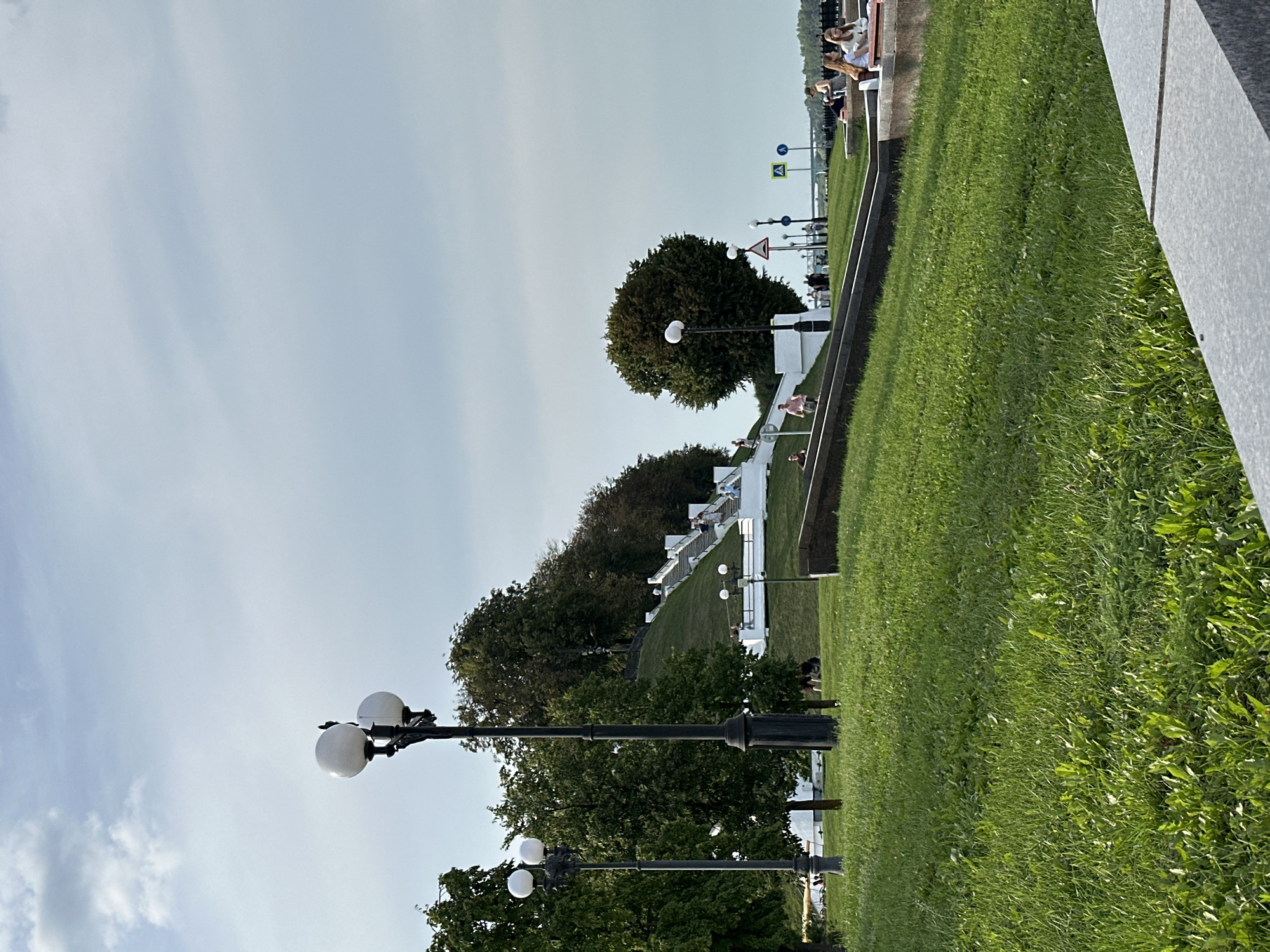 This is a ground-level, eye-level, medium shot of a park area with a large grassy slope leading up to a set of white stairs. The sky is overcast with scattered clouds. Two street lamps with white globes are visible in the foreground on the left side of the image. The grassy slope is well-maintained, and several trees are scattered throughout the park. A white staircase ascends the grassy slope, with several people walking up and down it. At the top of the stairs, a line of trees forms a treeline. In the distance, a body of water is visible, possibly a river, with a bridge crossing it. On the right side of the image, a group of people are sitting on benches, facing away from the camera. There are several traffic signs visible near the river. This scene appears to be in a public park within Rusanovo, Russia, a locale known for its scenic riverfront and recreational spaces.