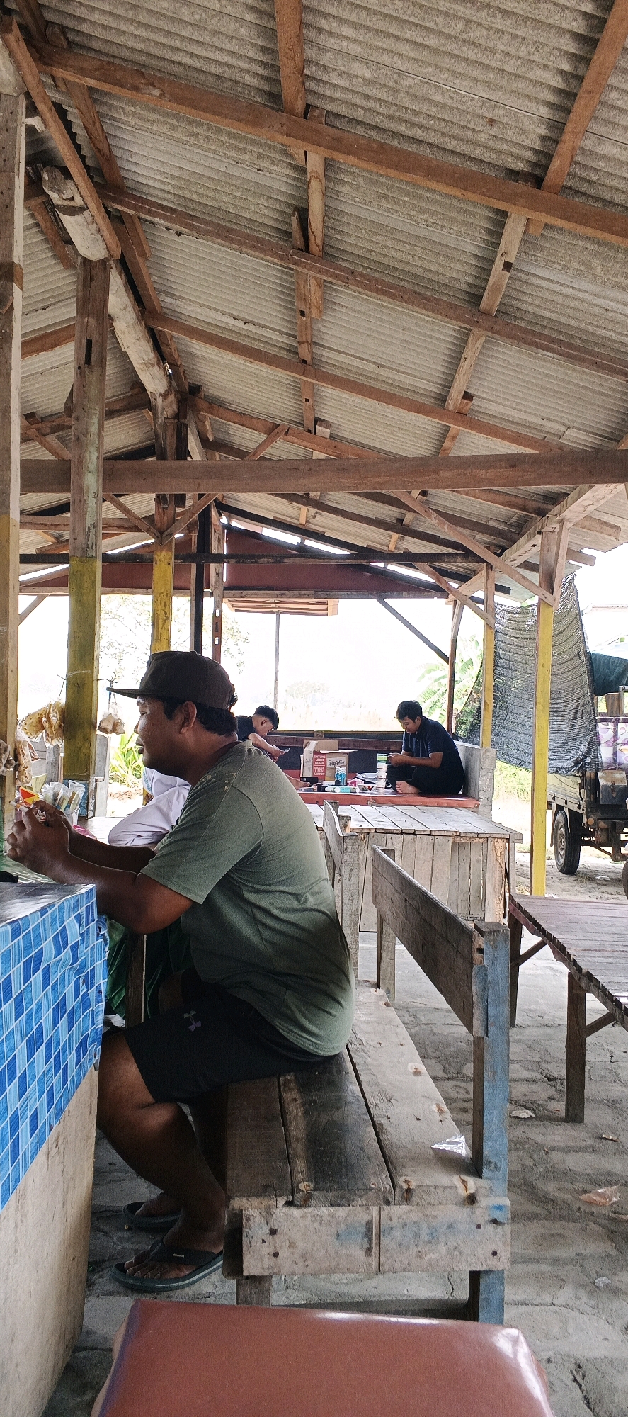 In Sonoadi, Indonesia, an outdoor seating area features a rustic wooden roof supported by posts. A man in a green t-shirt and dark shorts sits on a wooden bench, facing left, holding an object in his hands. In the background, another man in a dark shirt sits on a wooden platform, looking down at his phone, while a third person in a black shirt is also seated. The area appears to be a casual gathering spot, possibly a food stall or rest stop, with a wooden table and bench in the foreground and a truck visible in the distance.