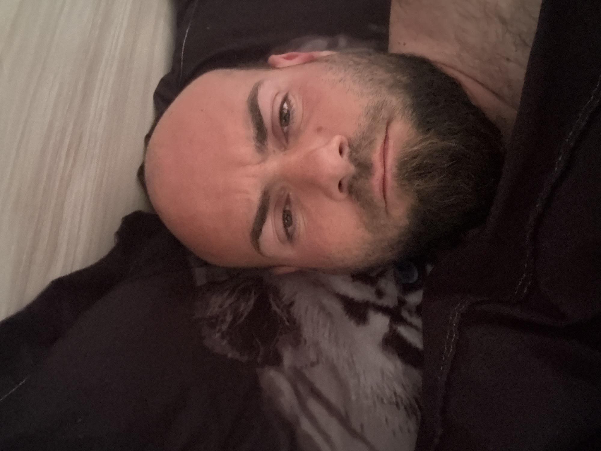 A man is lying down, appearing to be on a bed or similar surface. He has a shaved head with some stubble, a beard, and light-colored eyes. His face is slightly turned to the side. He is wearing a dark-colored garment. The bedding appears to be a combination of white and grey with a dark-colored cover. There is no information in the image to suggest he is in Sarnevo, Bulgaria.