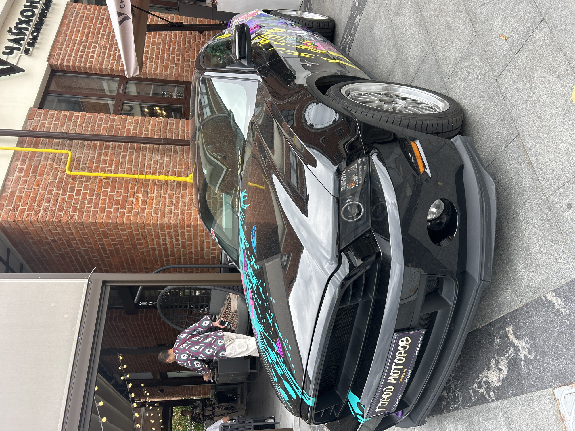 A black Ford Mustang with colorful paint splatters on its hood and side is parked on a stone-tiled sidewalk in front of a brick building. A sign on the car's license plate reads "ГОРОД МОТОРОВ." In the background, a person in a patterned shirt and light-colored pants walks past an open doorway. The building has large windows and a white awning extending from an upper level. The scene appears to be an outdoor dining area, possibly a cafe or restaurant, with tables and chairs visible. A yellow pipe runs vertically up the brick wall.