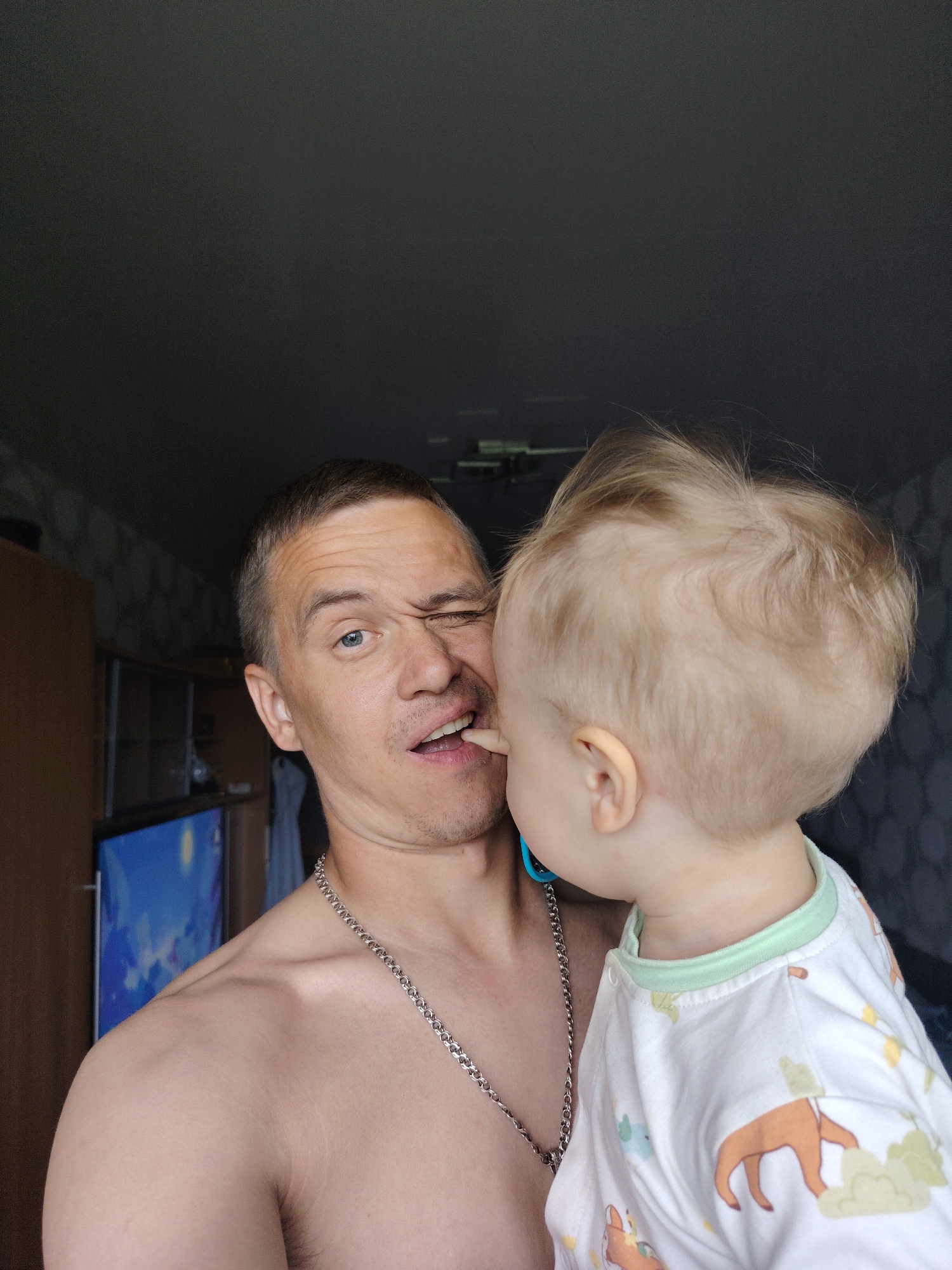 The image shows a shirtless man holding a toddler. The man is light-skinned with short brown hair and a silver chain necklace. He is winking and has his mouth slightly open with the toddler's finger inside. The toddler is light-skinned with blond hair, wearing a white onesie with animal designs and a light green trim. The toddler has a blue pacifier attached to their clothing. The background consists of dark walls and furniture. A TV is visible on the left. The location is identified as Chaikovsky, Russia.