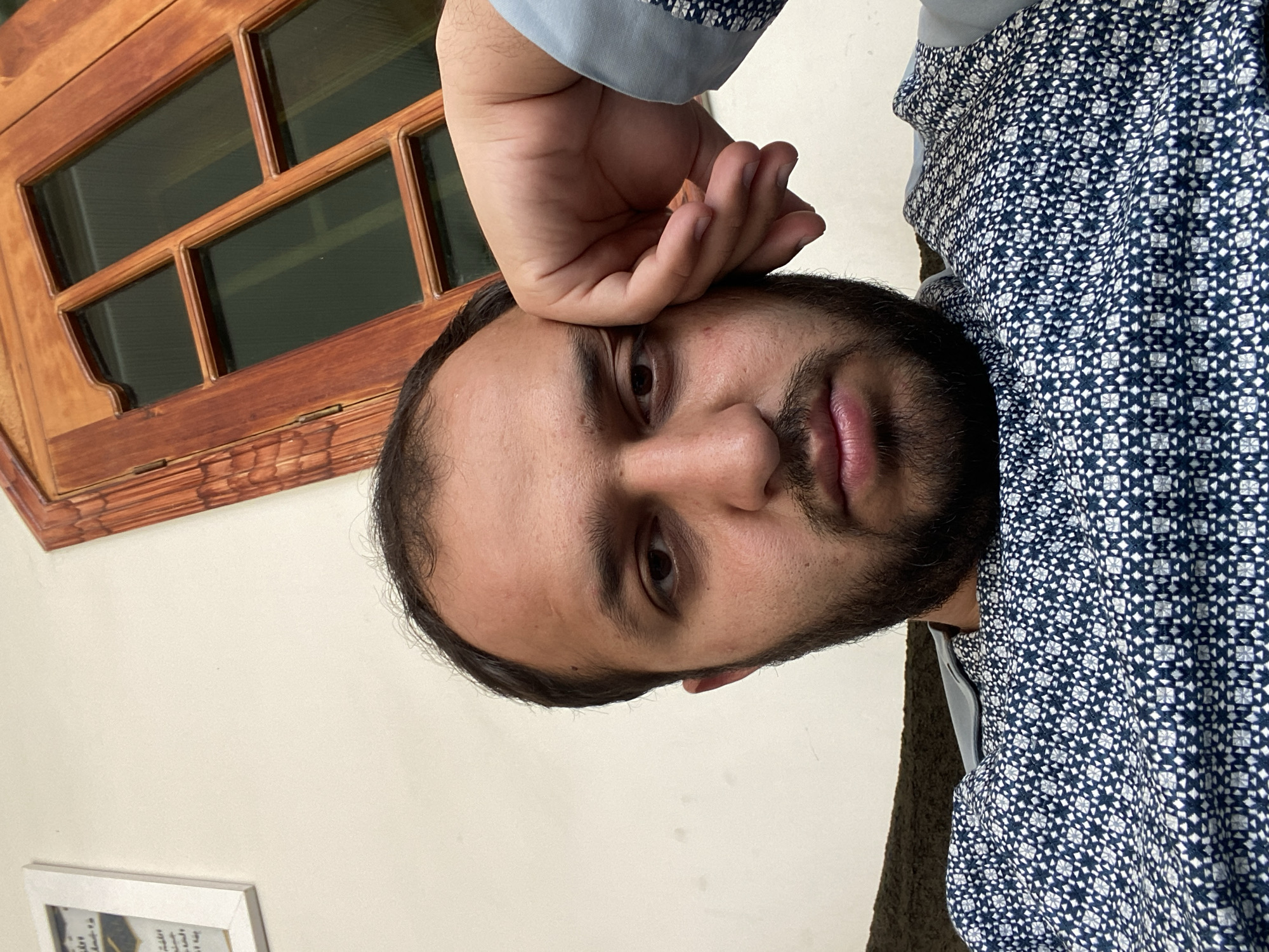 A man is captured in a close-up, selfie-style photograph, resting his forehead and hand on his face. He has a beard and is wearing a patterned shirt. The background suggests an indoor setting with a cream-colored wall. A framed picture is visible on the left, and a wooden decorative element, possibly a window frame or cabinet, is on the right. The overall lighting is soft, creating a slightly subdued atmosphere. The context of the image is not evident, but the visual elements present an ordinary domestic scene.