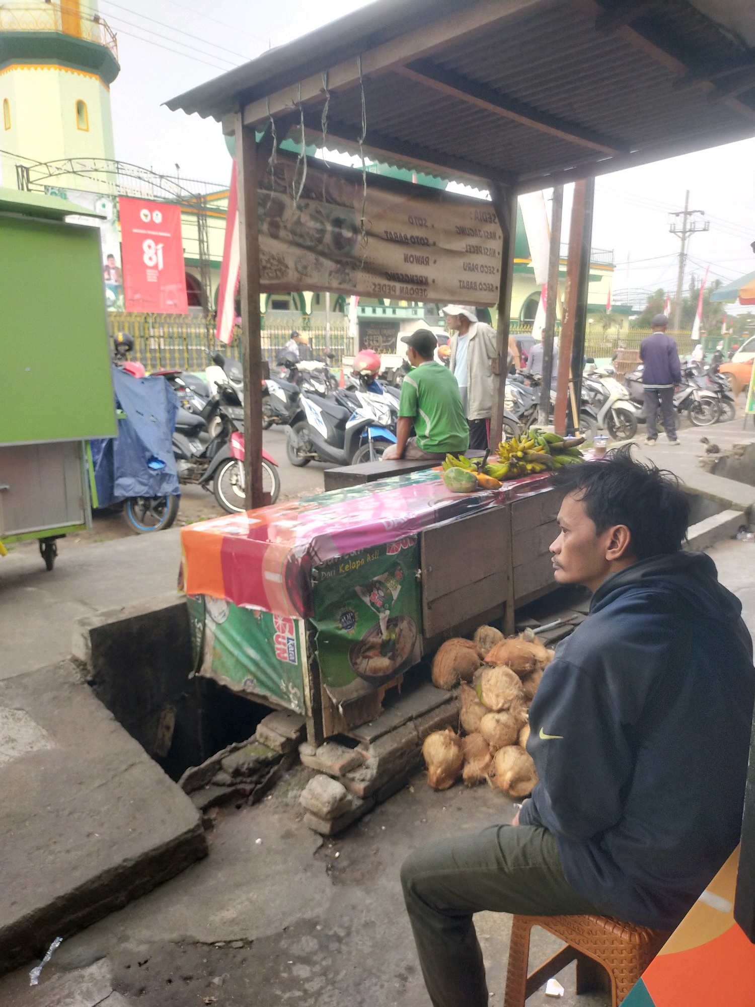 The image depicts a street scene in Bululawang, Indonesia, likely near a market or roadside stall. In the foreground, a young man with dark hair, wearing a dark blue hooded sweatshirt and olive green pants, sits on a woven orange plastic chair, facing left. He appears to be observing something to his left, with his right hand resting on his lap. Behind him, a wooden stall is visible, covered by a corrugated metal roof. The stall displays various items, including a pile of coconuts in front and a bunch of bananas and other fruits on top. Colorful banners are draped over the stall, one with the text "Dau Kelapa Asli" (Original Coconut Water) and another featuring a cartoon image of a soup bowl. A man in a white cap and light-colored jacket stands behind the stall, and another man in a green shirt sits on the stall's counter. Several motorcycles are parked behind the stall, and a blue tarp covers part of a green food cart. In the background, a mosque with a yellow and green minaret is visible, along with other buildings and a busy street with pedestrians and vehicles. The environment appears somewhat aged, with some areas of disrepair, particularly a broken section of concrete in the foreground. The sky is overcast.