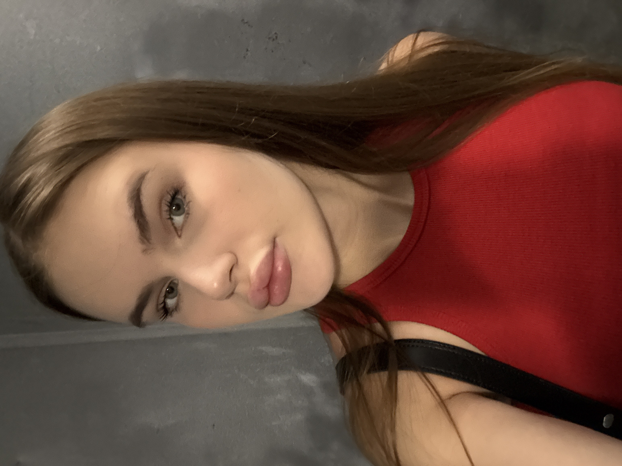 A young woman with long brown hair, fair skin, and green eyes is taking a selfie against a grey concrete wall. She is wearing a red ribbed tank top and a black strap is visible over her shoulder. Her lips appear to be enhanced. The image provides no information about her location in Dnipro, Ukraine, or any other details of the environment.