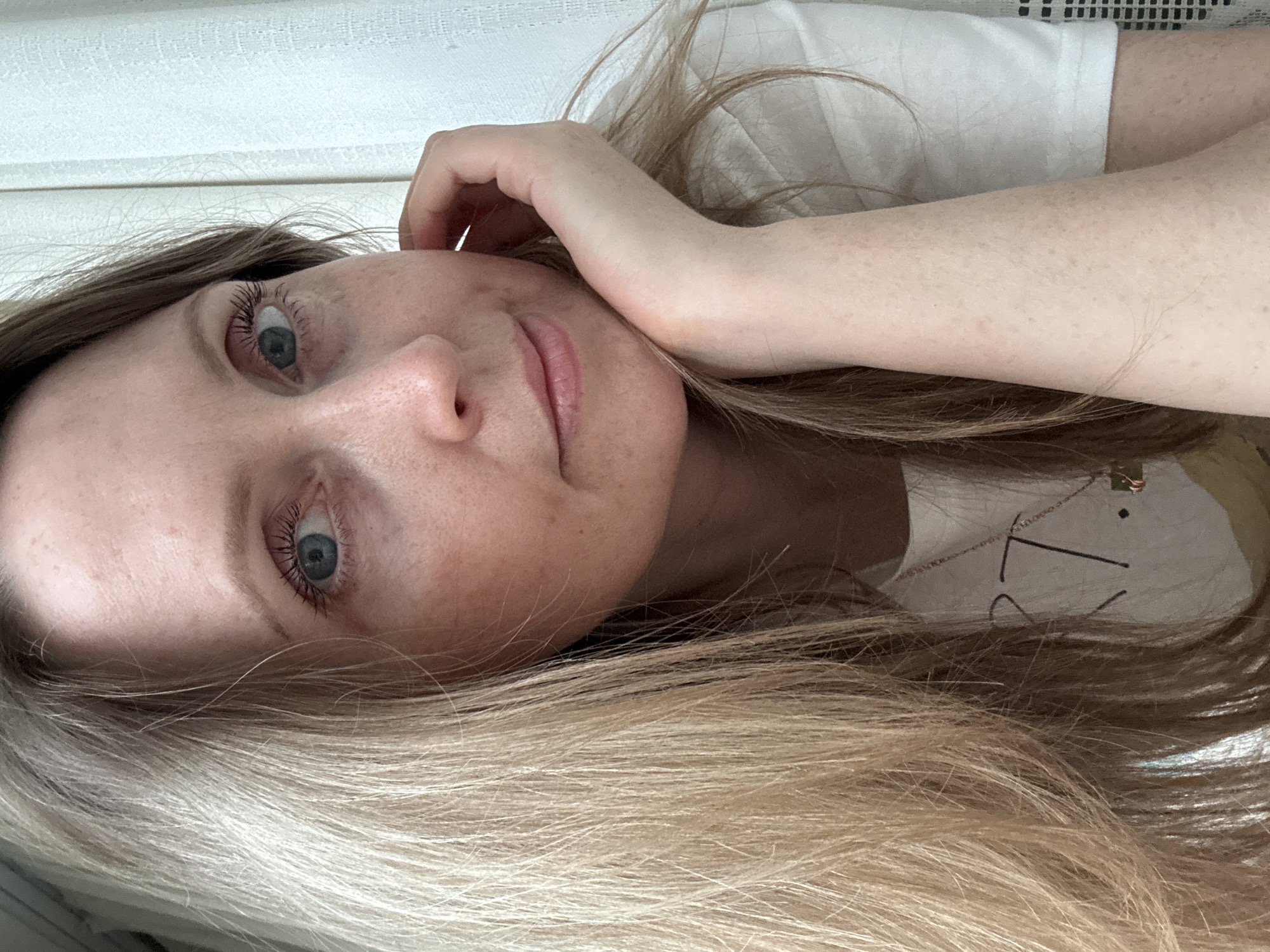 A woman with blue eyes and blonde hair lies on her side with her head resting on her hand. She is wearing a white t-shirt and a necklace. The background is a white wall. It is not possible to determine the location of the image.