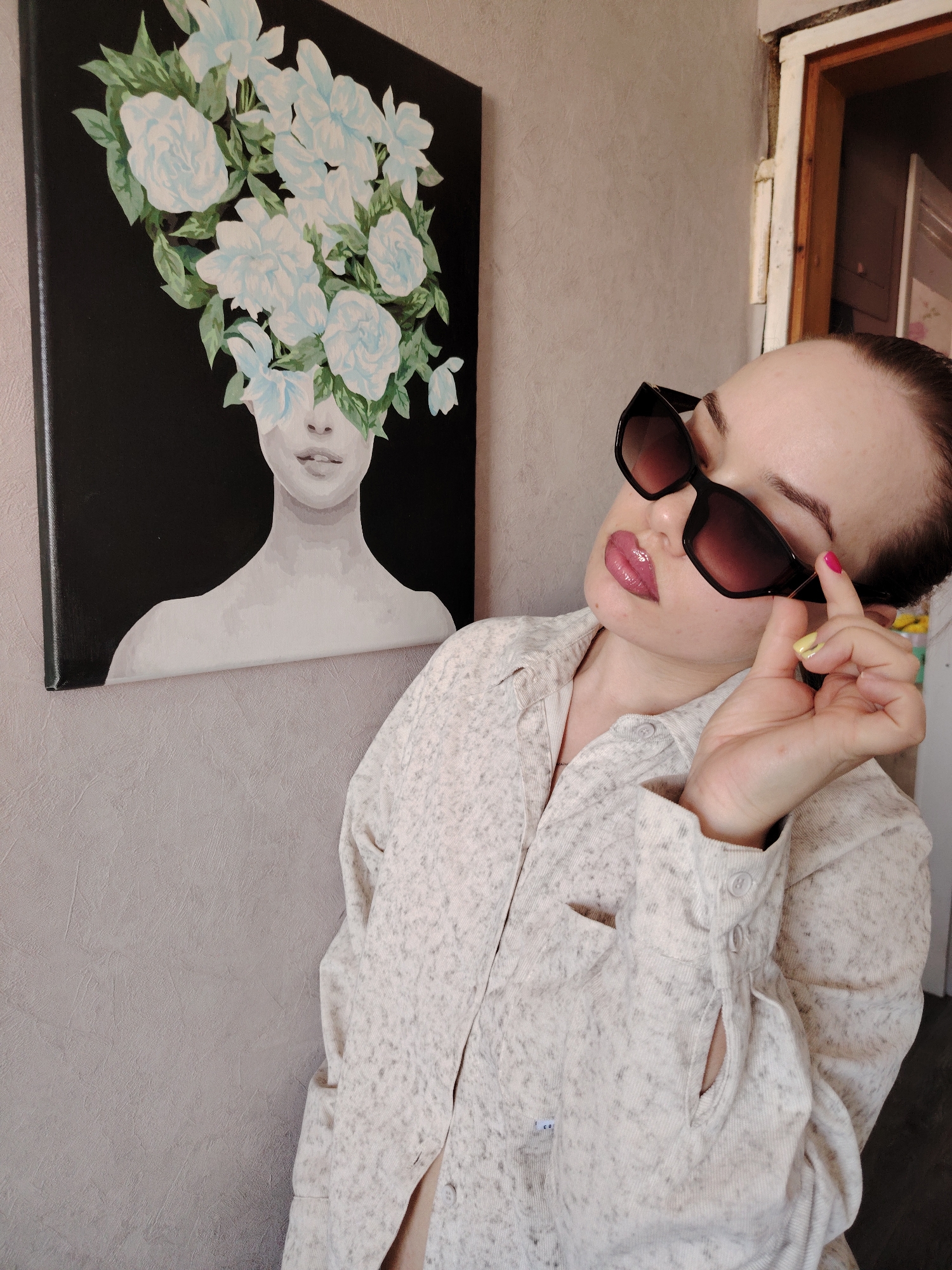 The scene is an interior location within the city of Марковский, Russia. A female subject is visible, angled slightly upward and to her left, with her right hand holding the right arm of dark, rectangular sunglasses. Her right index fingernail is painted bright pink, and her middle fingernail is yellow. Her lips are glossy pink. She wears a light-colored, speckled long-sleeved button-up shirt, partially unbuttoned at the collar. Behind the subject, a canvas painting hangs on a textured, light grey wall. The painting depicts the upper torso of a person with light blue and white flowers and green leaves obscuring the face, set against a black background. To the far right of the frame, a distressed wooden door frame is partially visible.
