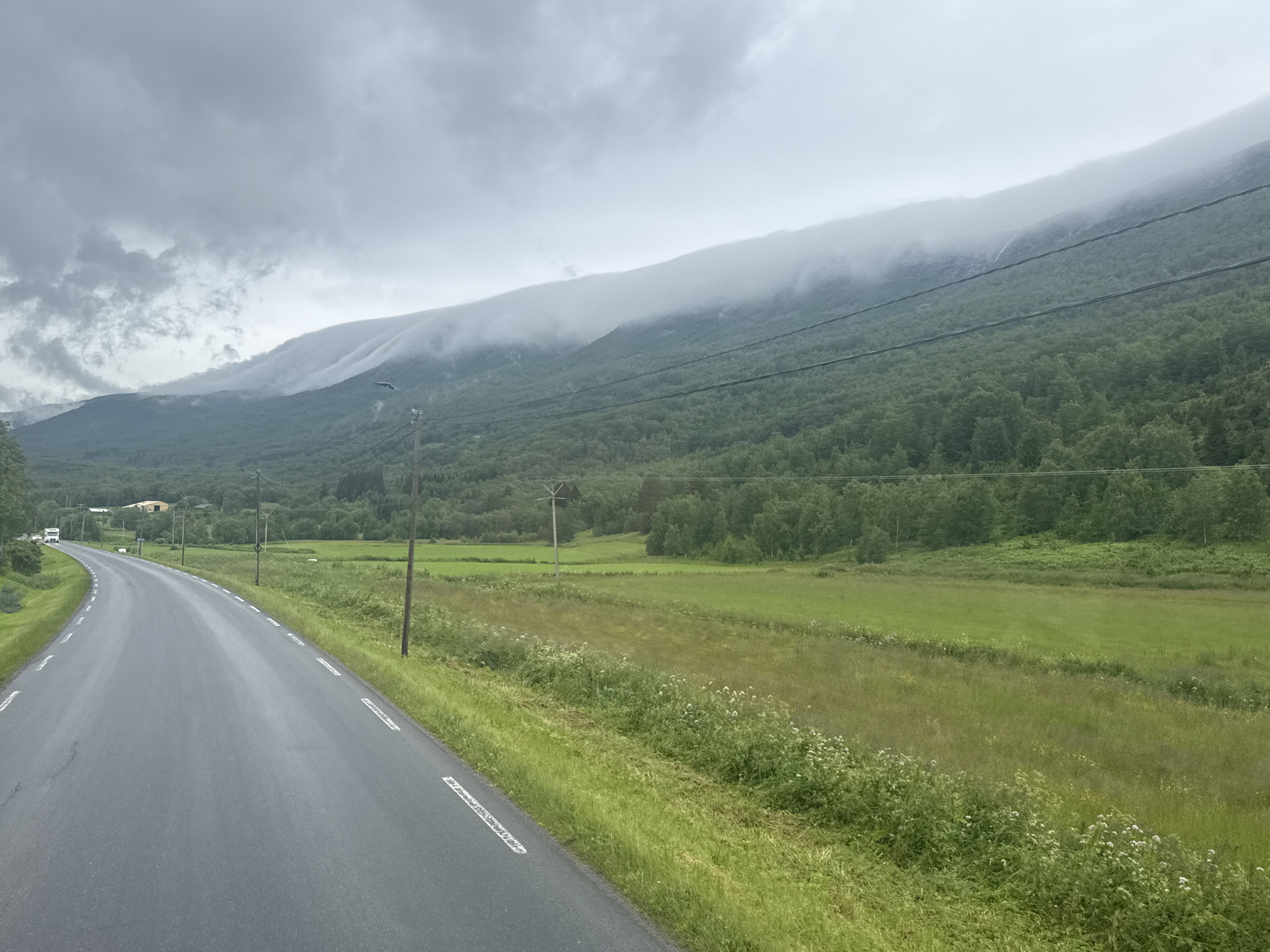 The image shows a road curving to the left, flanked by green grassy fields and a dense forest. In the background, a misty mountain range is visible, with the peaks obscured by clouds. The sky is overcast with a dull gray hue. A white line marking divides the lanes of the asphalt road. There are utility poles along the side of the road. The scene appears to be a rural or semi-rural landscape in Norway.