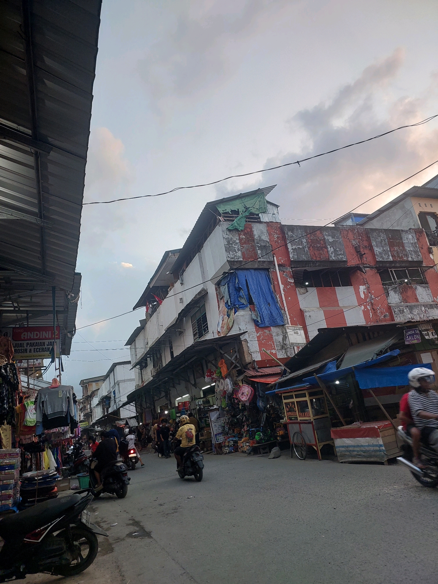 This is a full-shot, eye-level, vertical image of a street market scene in Ambon, Indonesia, likely during the late afternoon or early evening. The sky is overcast with clouds, hinting at impending or recent rain. The street is paved and appears to be a main thoroughfare in a bustling commercial district.
On the left side of the street, a covered market stall displays various clothing items, including a grey sweatshirt hanging prominently. A sign above the stall reads "TOKO ANDINI." Further down the street, more stalls and shops line the road, some with awnings providing shade.
The right side of the street features multi-story buildings with a mix of architectural styles and conditions. One building stands out with its weathered facade, painted with large red and white stripes, and draped with blue tarps. Other buildings are also visible, some appearing older and in need of repair. Small kiosks and food carts are set up along the sidewalk.
Several people are present, most of whom are on motorcycles. A person in a red and white striped shirt on a motorcycle is in the foreground on the right, moving away from the camera. A person wearing a yellow shirt and a yellow helmet is riding a motorcycle in the middle of the street, moving towards the camera. Other individuals on motorcycles are scattered throughout the scene, some appearing to be shoppers or vendors navigating the busy street. The overall impression is one of a lively, everyday commercial environment in an Indonesian city.
