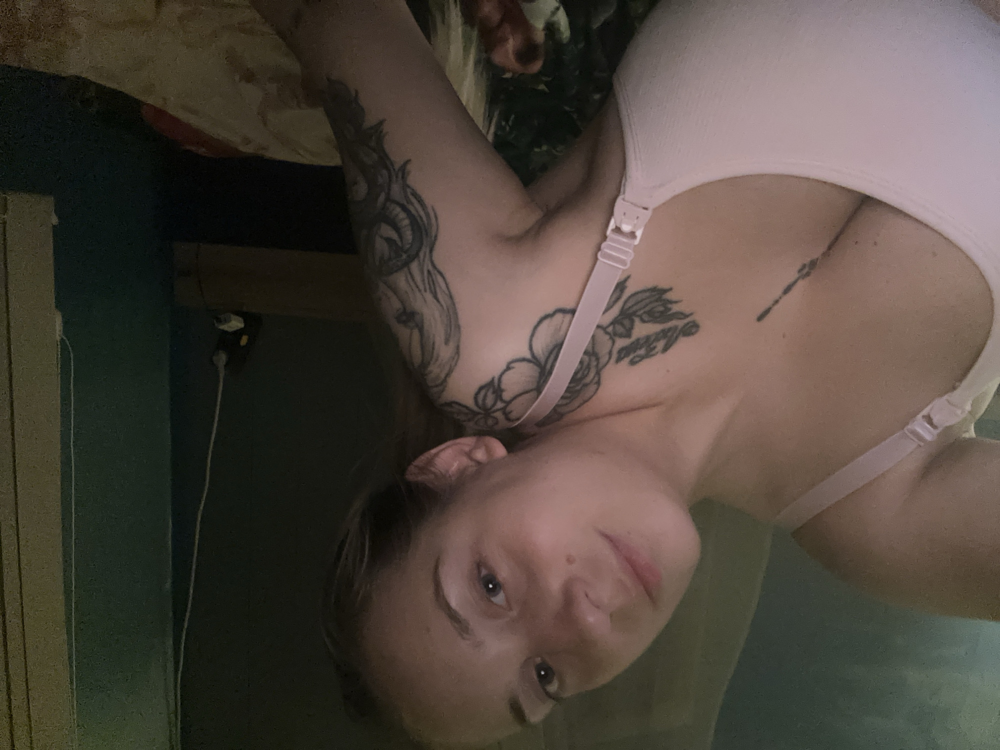 The image shows a close-up, slightly angled, selfie of a young woman with light skin, brown eyes, and blonde hair. She is wearing a light pink bra and has multiple tattoos visible on her chest and left arm. Her head is tilted back, and she is looking towards the camera. The background appears to be an indoor setting with dark walls and a bed. There are no discernible Plovdiv, Bulgaria specific elements in the image.