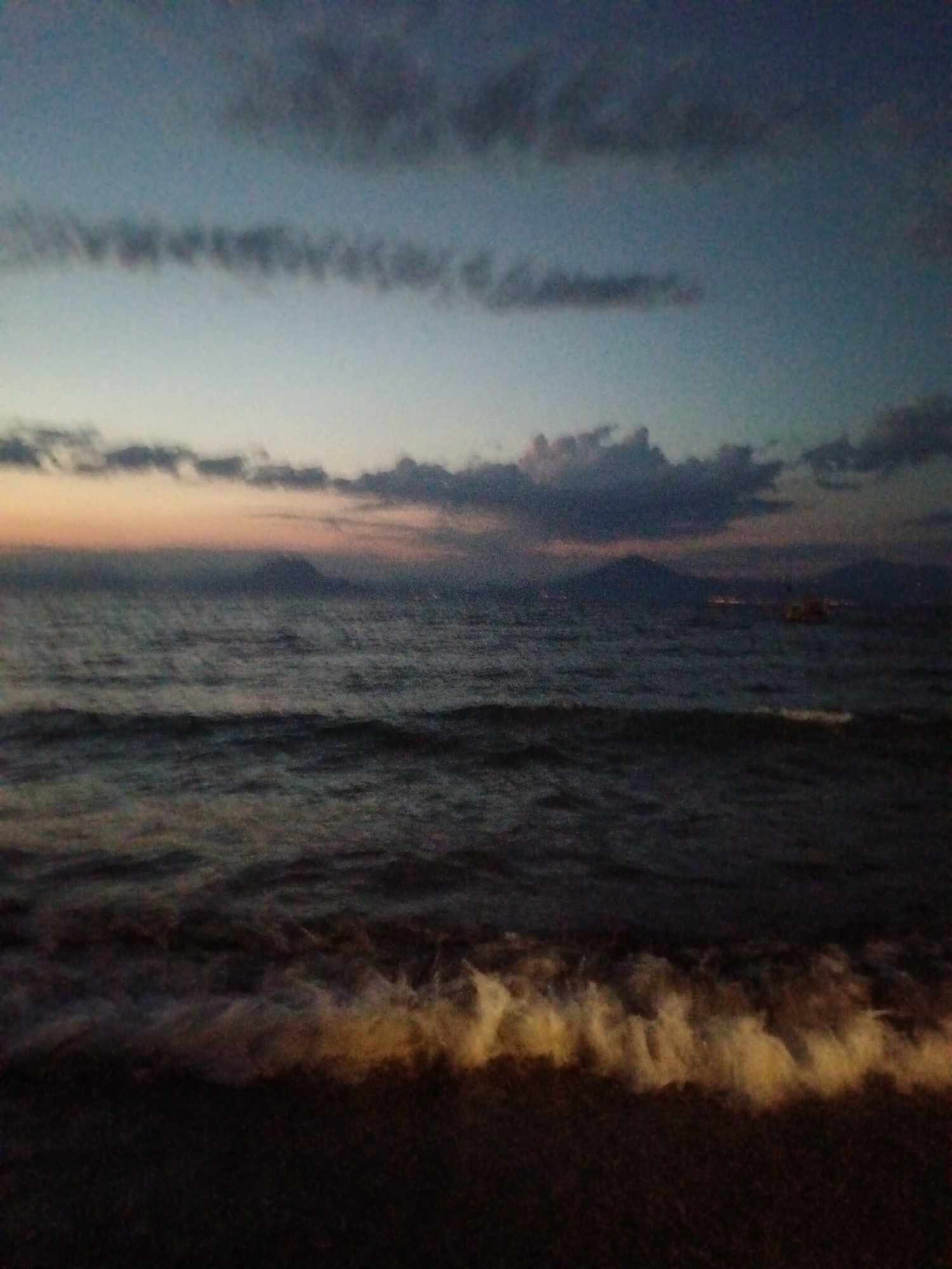 The image depicts a twilight scene at a beach in the Municipal Unit of Vrachneika, Greece. The sky is a gradient of deep blue at the top, transitioning to lighter blues and a band of orange and pink near the horizon, indicating sunset or sunrise. Several clouds are visible, some dark and heavy, others wispy and elongated. The sea is dark and choppy, with white foam from waves crashing on the shore. The foreground is a dark beach area, with the white froth of the waves being the brightest element. In the distance, silhouetted landforms, possibly mountains or islands, are faintly visible against the horizon, with some scattered lights suggesting a settlement. There is a small, indistinct boat or vessel in the water on the right side of the frame. The overall impression is one of a serene, natural environment at the edge of day. There are no people or discernible human activity in the scene.
