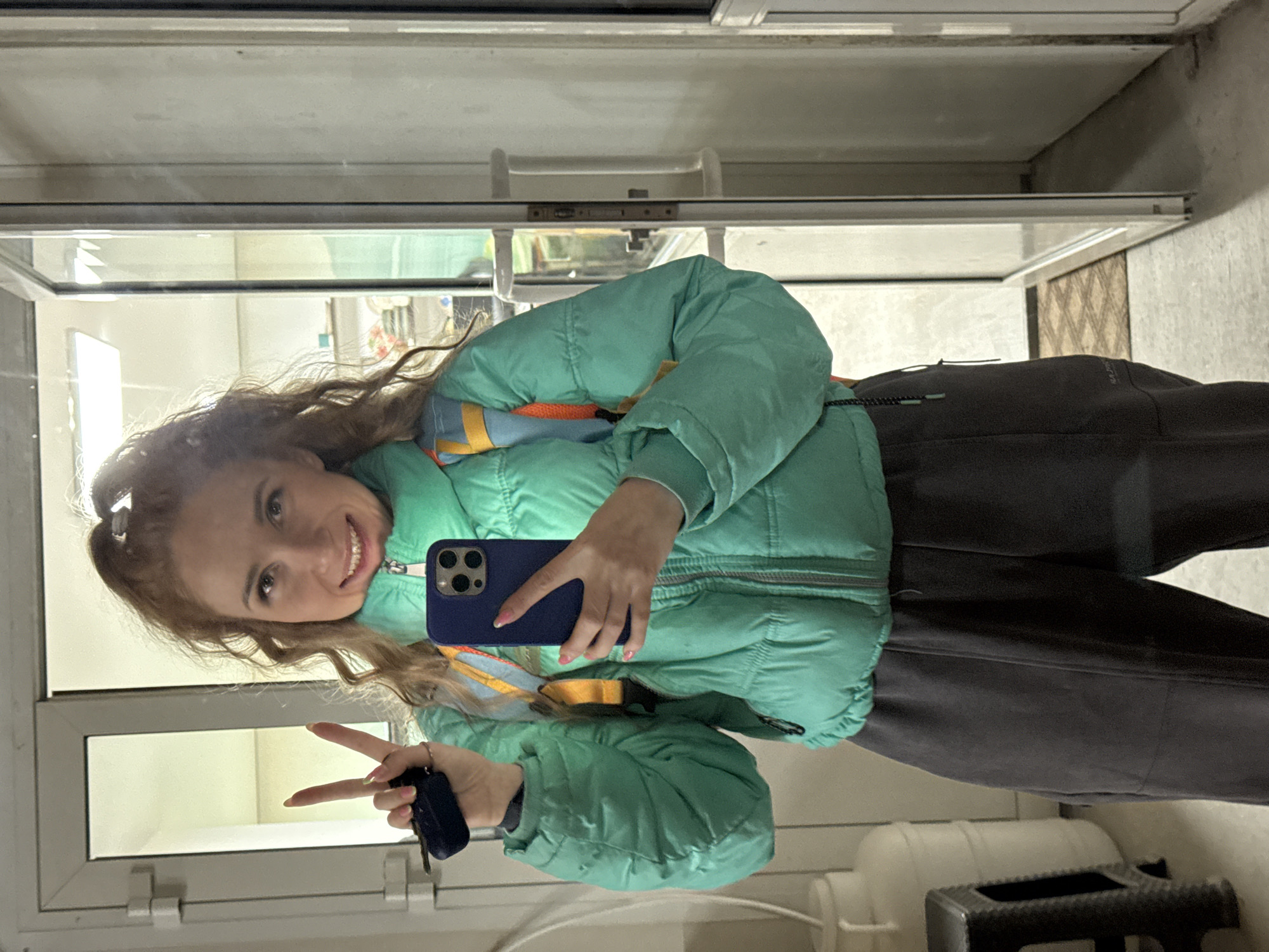 A young woman with long curly brown hair is taking a selfie in front of a glass door. She is wearing a light green puffer jacket and dark pants. She is holding a blue smartphone in her right hand and making a peace sign with her left hand, which also holds a black key fob. The reflection in the glass door suggests that the location is inside a building. The location is in Chaikovsky, Russia.