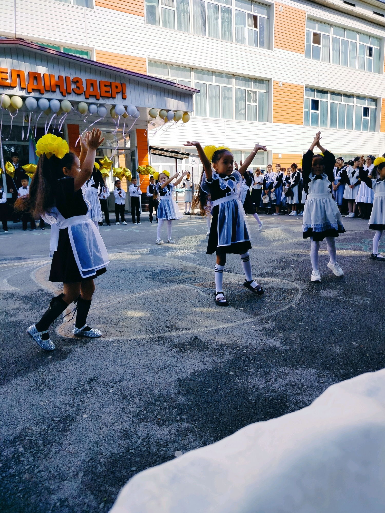 In front of a modern, multi-story building with beige and white siding, a group of young girls in school uniforms are celebrating outdoors. The girls are dressed in traditional black dresses with white pinafores and bows, some with yellow hair accessories. Several girls are dancing with their arms raised, while others stand in a line behind them. In the background, balloons and star-shaped decorations are visible. The building has large windows and a prominent orange sign above the entrance with text in an unfamiliar script, likely a school or official building in the city of Kölkent, Kazakhstan. The ground is asphalt, marked with white lines. The scene appears to be a school event, possibly a celebration or graduation ceremony.
