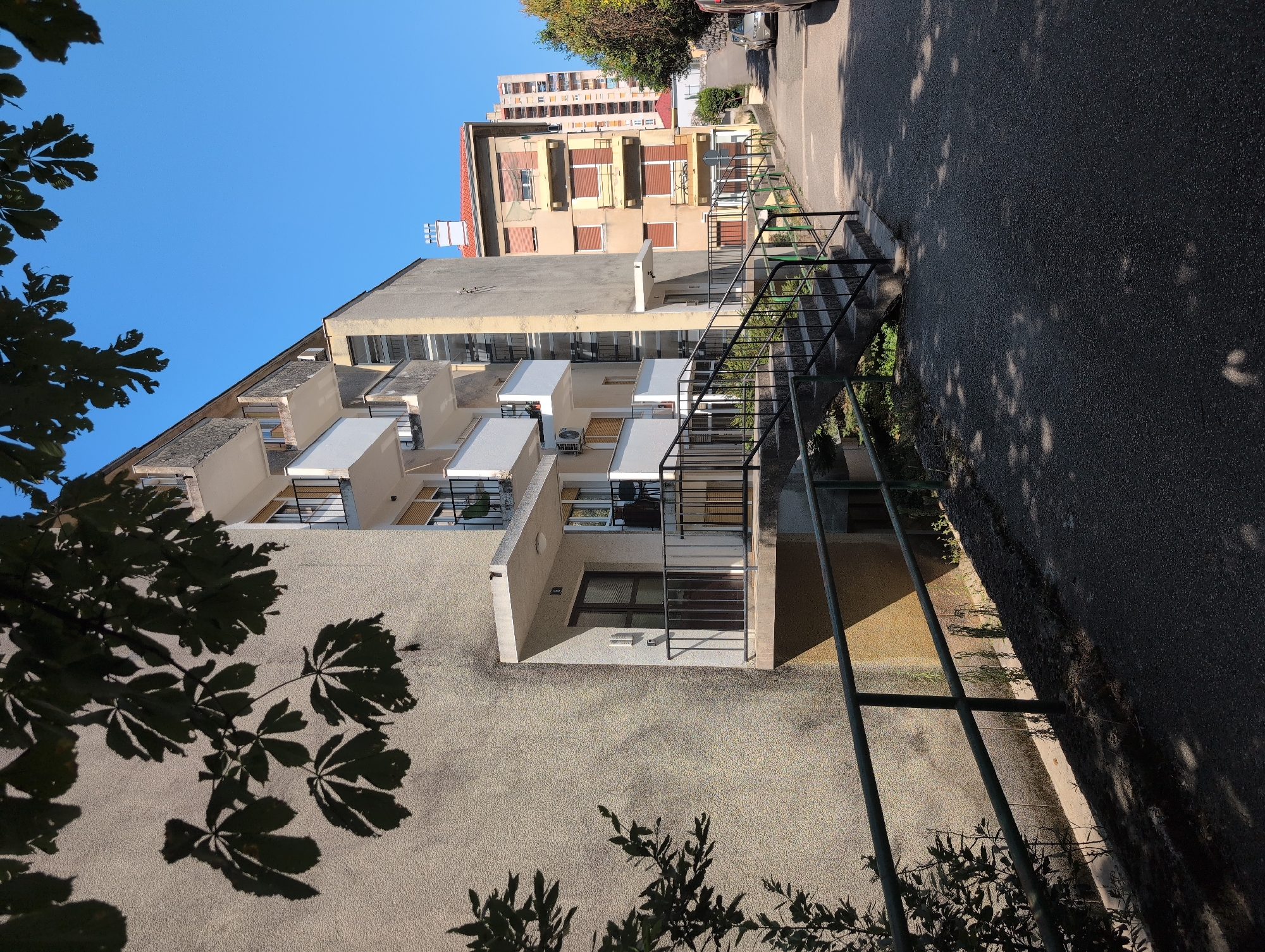 The image captures a street scene in the city of Rijeka, Croatia, featuring residential buildings and a clear blue sky. In the foreground, a paved road runs diagonally from the bottom right to the middle distance. A dark green metal railing runs along the left side of the road, delineating a raised embankment. Above this embankment, the textured, light beige wall of a building is visible, with shadows from tree leaves cast upon it.
To the right of the embankment, a concrete staircase with dark metal railings ascends towards the buildings. The first building on the left is a multi-story structure with prominent white balconies and gray textured walls. The entrances to apartments are visible, with dark doors and small windows. Above one of the doors, the number "7" is displayed.
Further down the street, another building, painted in shades of beige and yellow, stands with windows covered by brown shutters. A bicycle is visible leaning against this building. In the background, taller, more modern apartment buildings are seen, with a lighter facade. The scene is bathed in sunlight, suggesting a daytime setting. A silver car is partially visible on the right side of the road in the distance.