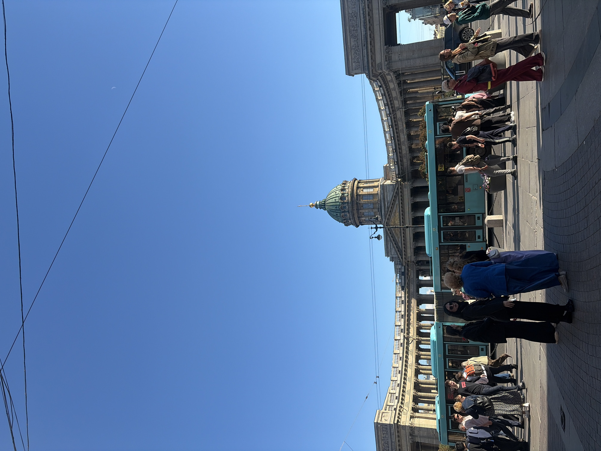 In a wide-angle shot of a clear blue sky with a faint crescent moon visible, a large, curved building with classical architecture dominates the lower half of the frame. The building features numerous columns, a grand semicircular colonnade, and a prominent green dome with a golden spire. In front of the building, a turquoise bus with a "24" visible on its side is parked, and a group of pedestrians are walking on a paved area. The people are casually dressed, some walking individually and others in small groups, with diverse ages and genders represented. The sun casts shadows, indicating it is daytime. The scene appears to be a public square or street in a city, with the architectural style suggesting a European location, potentially St. Petersburg, Russia, given the grandeur and the dome characteristic of Russian Orthodox cathedrals.