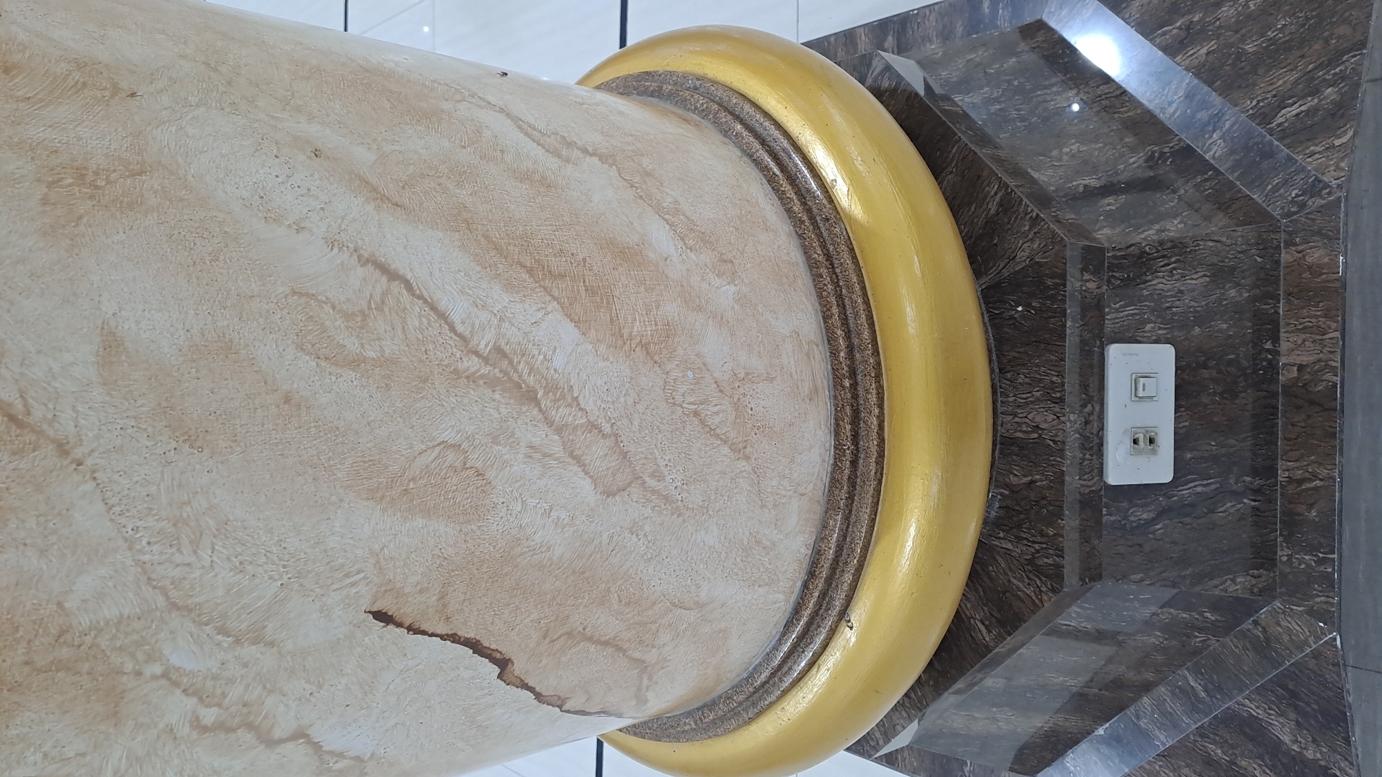 The image shows a close-up of a decorative column in what appears to be an indoor setting, possibly a public building or a well-decorated residence. The column is large and cylindrical, with a textured surface that mimics marble or a similar stone. It has a light beige or cream color with swirling patterns of brown and tan. A dark, irregular crack or imperfection is visible on the lower left portion of the column. The column is accented by a thick, golden band that wraps around it, creating a prominent decorative element. This band has a smooth, glossy finish.
Behind the column, the wall features a dark, marble-like pattern with a geometric design. The pattern is made up of angled, beveled surfaces that create a sense of depth and angularity. A white electrical outlet and light switch plate are mounted on this wall, positioned to the right of the column. The outlet has two sockets, and the switch appears to be in the "off" position. The lighting in the scene is bright, suggesting it is daytime or the area is well-lit artificially. There are reflections visible on the patterned wall, indicating a smooth, possibly glossy surface. No people or animals are present in the image, and no specific activity or event is depicted. The overall impression is one of a luxurious or well-appointed interior space with architectural embellishments. The presence of the electrical outlet suggests it is a functional space. There is no visible text in the image. The location context provided is Lhokawe Teungoh, Indonesia, which adds a regional aspect to the description.
