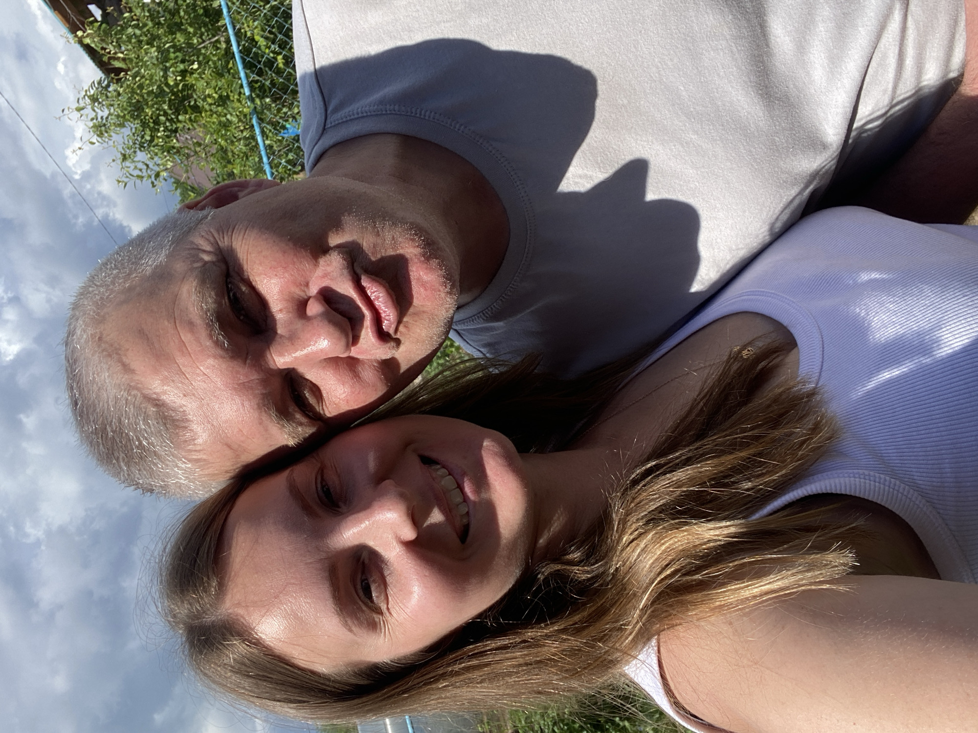 A close-up, selfie-style photograph captures a Caucasian man and woman in what appears to be a backyard or rural setting, likely on a sunny day with scattered clouds in the sky. The man, on the right side of the frame, has short, graying hair and is making a puckering expression with his lips. He is wearing a light gray t-shirt. The woman, on the left, has long, brown hair and is smiling broadly, looking towards the camera. She is wearing a white tank top. They are positioned closely together, with the man's head tilted towards the woman's. In the background, a chain-link fence with blue accents and lush green foliage are visible, suggesting an outdoor environment. The lighting indicates the sun is high in the sky, casting shadows. There is no visible indication of the city of Chaikovsky, Russia in the image itself.