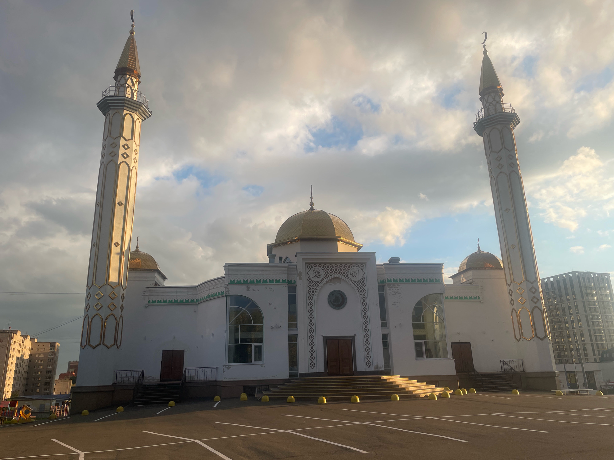 The image shows the "Idris" Mosque in Izhevsk, Russia, during the daytime. The mosque is a white building with a golden dome and two tall white minarets topped with crescents. The minarets have decorative golden patterns. The mosque has an entrance with a set of stairs leading up to double wooden doors. Large arched windows are visible on the sides of the mosque. The foreground features a paved parking lot with white markings. In the background, to the left, are several multi-story residential buildings and a playground with a yellow slide. To the right of the mosque, a modern, tall, glass-fronted building is visible. The sky is cloudy with patches of blue. No people are visible in the scene.