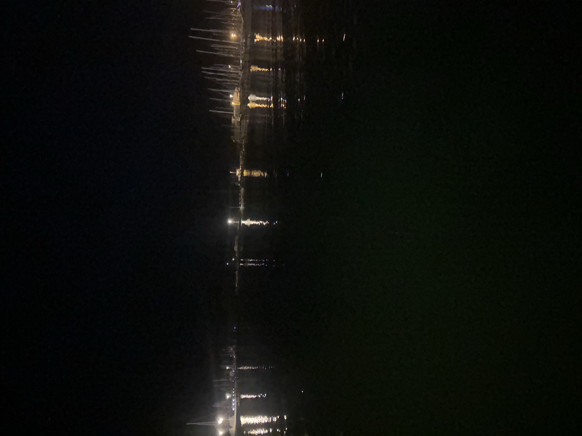 This is a nighttime, eye-level, full shot of a marina in Agde, France. The water in the foreground is dark and reflects the lights of the docked boats. In the background, a dense cluster of sailboats are moored, their masts creating a vertical line against the dark sky. Lights from buildings and street lamps illuminate the marina, casting long reflections on the water. The scene is quiet and still, suggesting a peaceful, serene atmosphere.