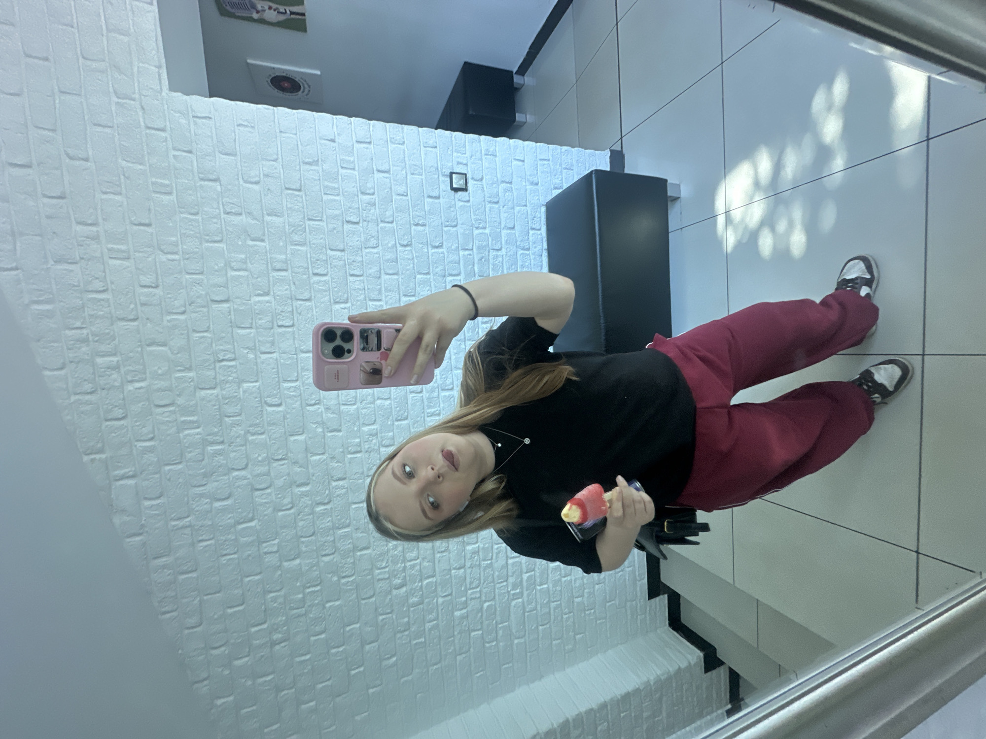 A young woman is taking a selfie in a room with white brick walls. She is wearing a black long-sleeved shirt and red sweatpants. She is holding a pink mobile phone in her left hand and an ice cream bar in her right hand. She is standing on a white tile floor next to a black bench. The location is Chaikovsky, Russia.