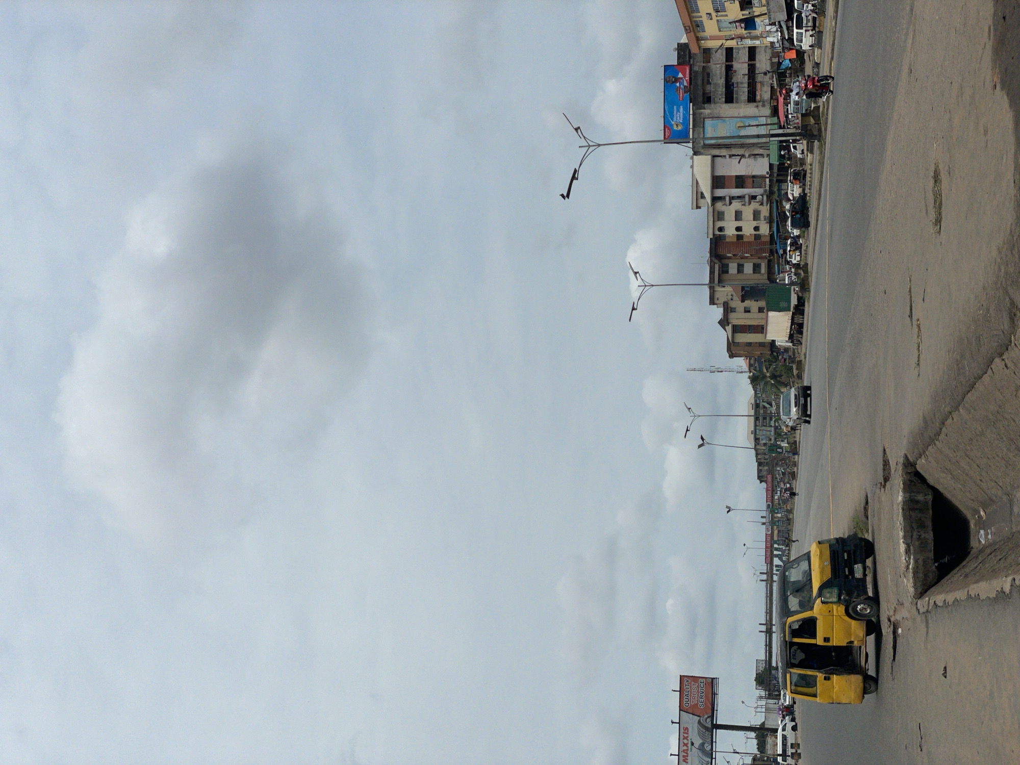 A street scene in Onitsha, Nigeria, featuring a road with a yellow minibus parked near an open drain. Buildings line the street, and a billboard is visible in the foreground. The sky is overcast. Other cars and people are visible along the street. Street lights are present.