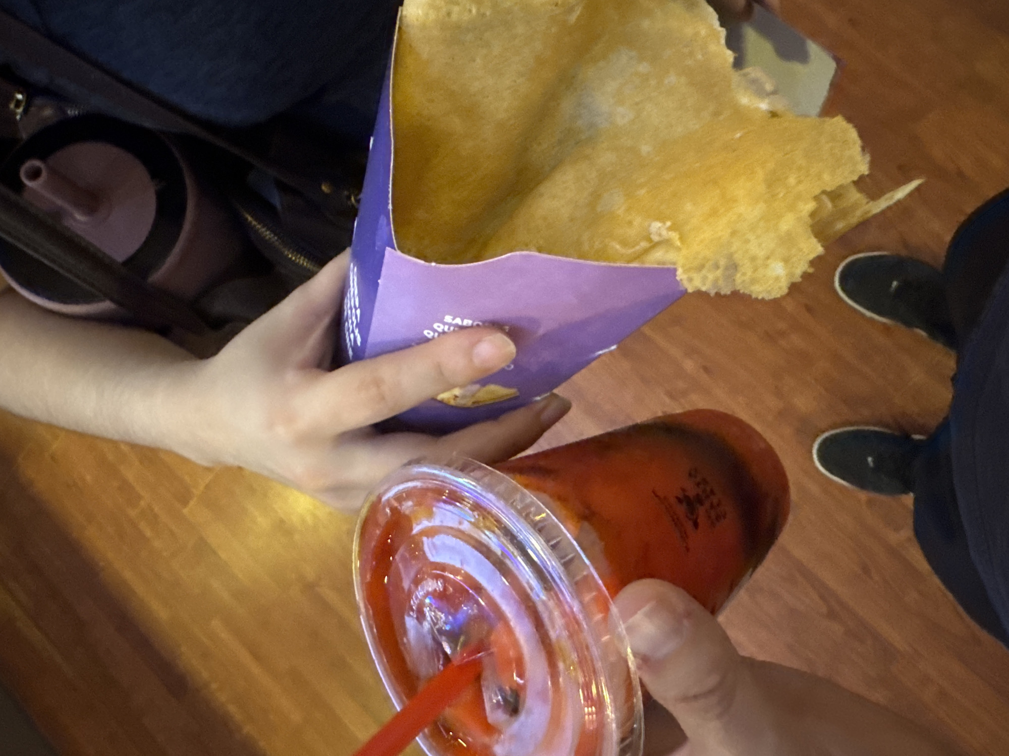 A high-angle shot depicts a person holding a crêpe in a purple wrapper in their left hand and a beverage in a clear plastic cup with a red straw in their right hand. The crêpe appears to be filled and golden in color. The drink is reddish. Part of a handbag with a pink tumbler inside is visible to the left of the crêpe, as are the feet of another person wearing dark shoes and pants. The background is a wood-look floor. The location is Puebla, Mexico.