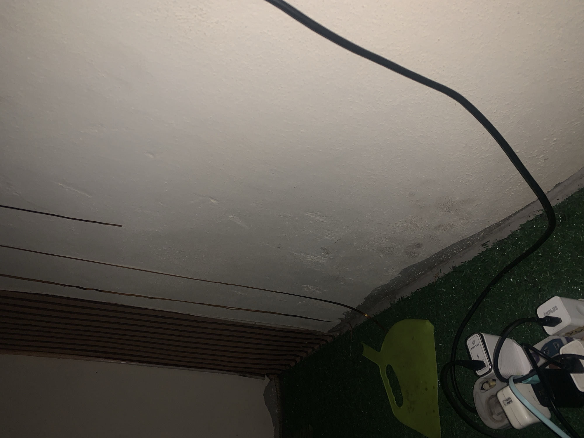 The image depicts a close-up, low-angle view of the interior corner of a room. The floor is covered with artificial turf, and a lime green dustpan lies on it. In the foreground, several electrical adapters and extension cords are plugged into each other, creating a tangled arrangement. The wall is painted white, with some apparent discoloration and texture variations. To the left, a portion of a slatted wooden paneling is visible, running vertically. Several thin wires or cables are seen running vertically along the wall. The lighting is dim, suggesting an indoor setting with artificial light. The scene is devoid of people and any overt actions. The location is specified as Jalingo, Nigeria.