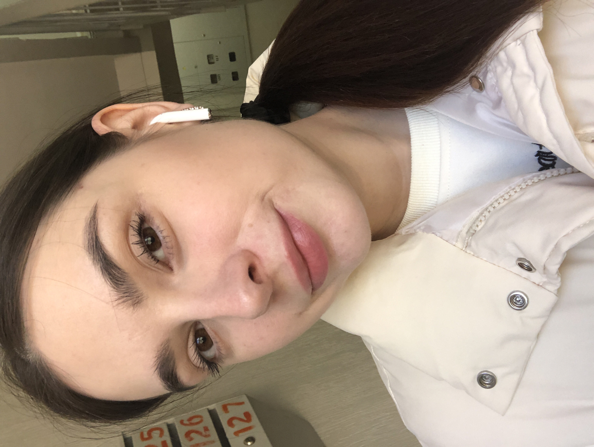 A medium close-up photo shows a woman from the chest up, rotated clockwise approximately 90 degrees. Her dark brown hair is pulled back and tied. She is wearing a white crew-neck shirt and a white puffer jacket. An earbud is in her left ear. In the foreground is a light-colored wooden surface. In the lower left corner are white and orange markers with numbers. The numbers 25, 126, and 127 are visible. In the background is a blurry image of what appears to be a white wall with control panels. There is no evidence in the image to suggest that it was taken in Chaikovsky, Russia.