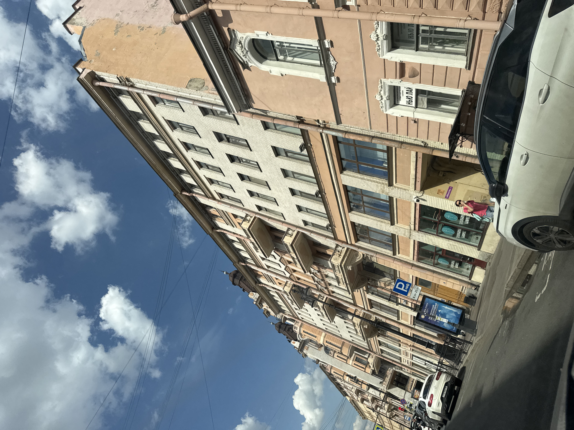 The image presents a vibrant, tilted street scene in Saint Petersburg, Russia, on a bright, sunny day, likely captured from a moving vehicle. Multi-story buildings line the street, featuring classic European architecture with pastel-colored facades, ornate window frames, decorative balconies, and visible drainpipes. Some buildings exhibit signs of age with peeling paint and varying textures, such as brick and smooth plaster.
A brilliant blue sky, adorned with scattered white cumulus clouds, dominates the upper left portion of the frame, with thin power lines crisscrossing across it. Several cars are parked along the curb, including a white SUV that partially obscures the right side of the image and another white car further down the street.
A person wearing a pink top and dark trousers is visible on the sidewalk near a building entrance. Street elements include a blue parking sign displaying "P 8.00-20.00" and a digital advertising screen showcasing an image of a building with partial Cyrillic text reading "ПЕТЕРБУРГСКИЙ НАРОДНЫЙ". Other visible text on storefronts includes "НЬЮ ЛАЙ" (New Line) and "СПЕЦИАЛИЗИРОВАННЫЙ САЛОН" (Specialized Salon). A small white sign in a window prominently displays "ROSTIC'S".