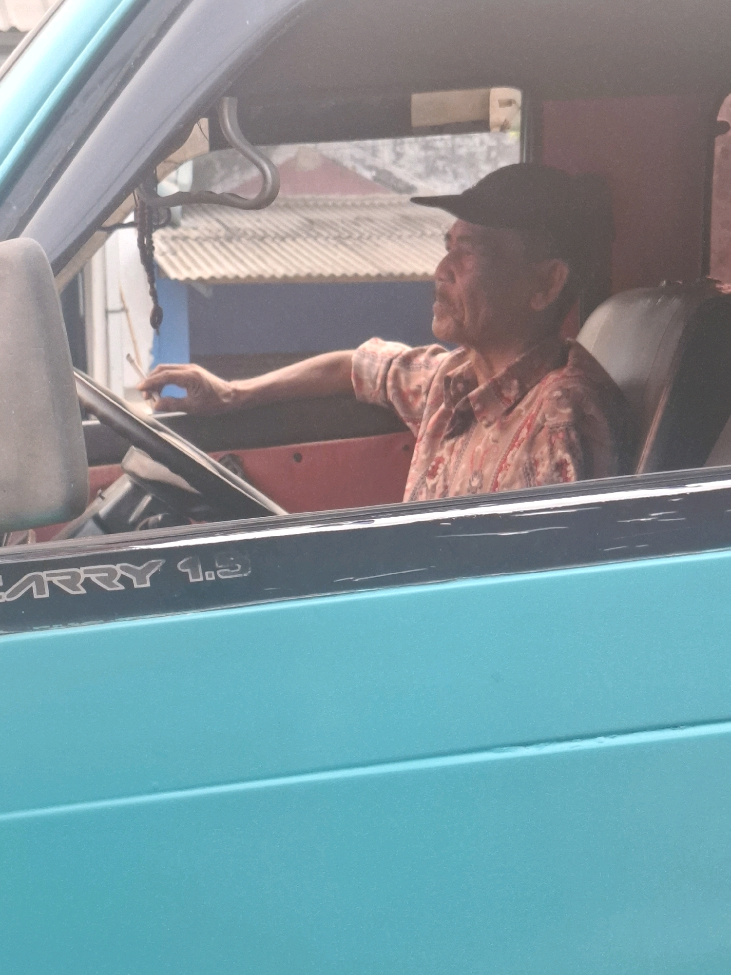 In Cijalingan, Indonesia, an elderly man with a cap is seated in the driver's seat of a turquoise vehicle. He is wearing a patterned shirt and is holding a cigarette in his right hand. The man is looking out of the vehicle's window. The vehicle's interior includes a steering wheel and part of a seat. Outside the vehicle, there are buildings with corrugated roofs. The phrase "CARRY 1.5" is visible on the side of the vehicle.