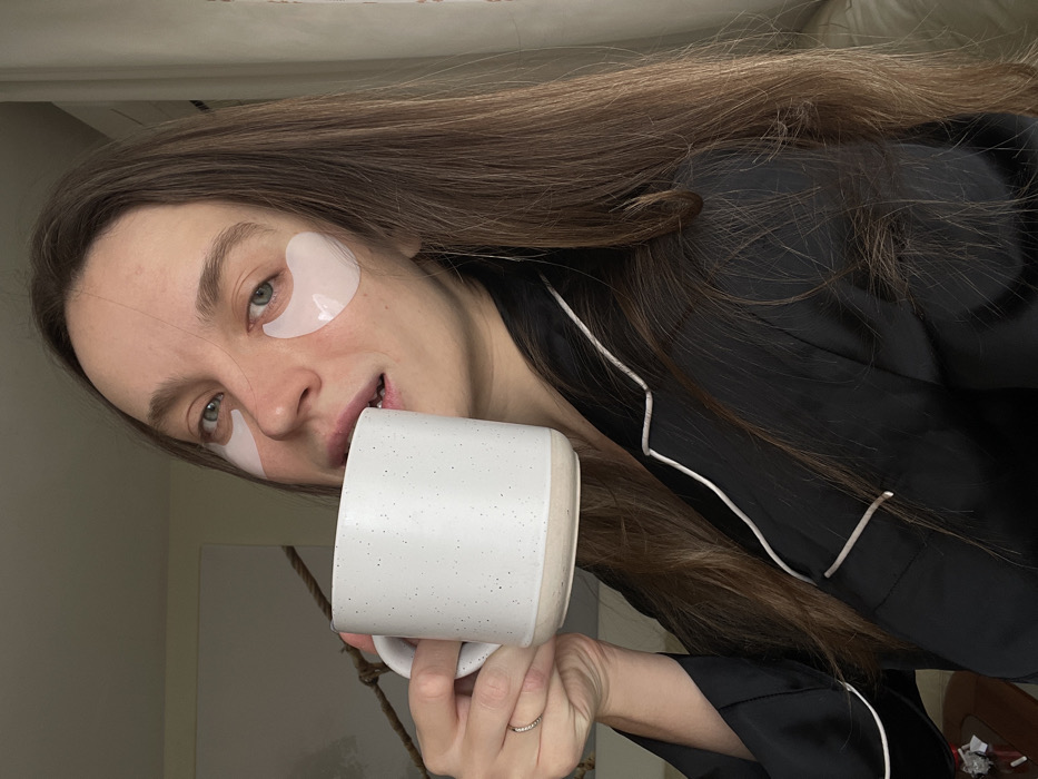 An indoor scene features a female subject positioned centrally. She has long, dark brown hair and light skin. Two translucent, light-colored eye patches are applied beneath her eyes. She wears a black long-sleeved top or pajama shirt with white piping along the collar and front opening. Her right hand holds a white ceramic mug, speckled with dark dots, to her lips; the mug has an unglazed, light brown base. A thin, silver-colored ring with small stones is visible on her right ring finger. She is looking directly towards the camera. The background comprises a light-colored wall, an indistinct dark rope or cable element in the upper left, and a partial view of light-colored furniture or fabric to the right. No visual identifiers within the image confirm the location as Budva, Montenegro.