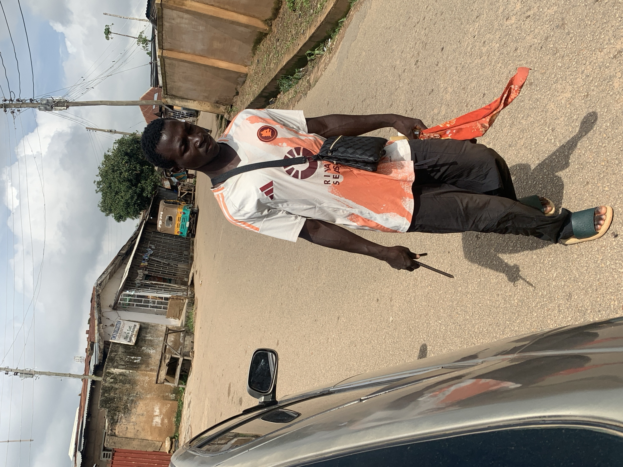 In Jos, Nigeria, a man walks down a paved street. He is wearing a white t-shirt with orange accents and a black crossbody bag. He holds a red cloth with a floral pattern in his right hand and a dark object, possibly a stick, in his left. The street is bordered by buildings and a concrete wall. A car is parked on the left side of the image. In the background, a yellow and green tricycle is visible. The sky is cloudy.
