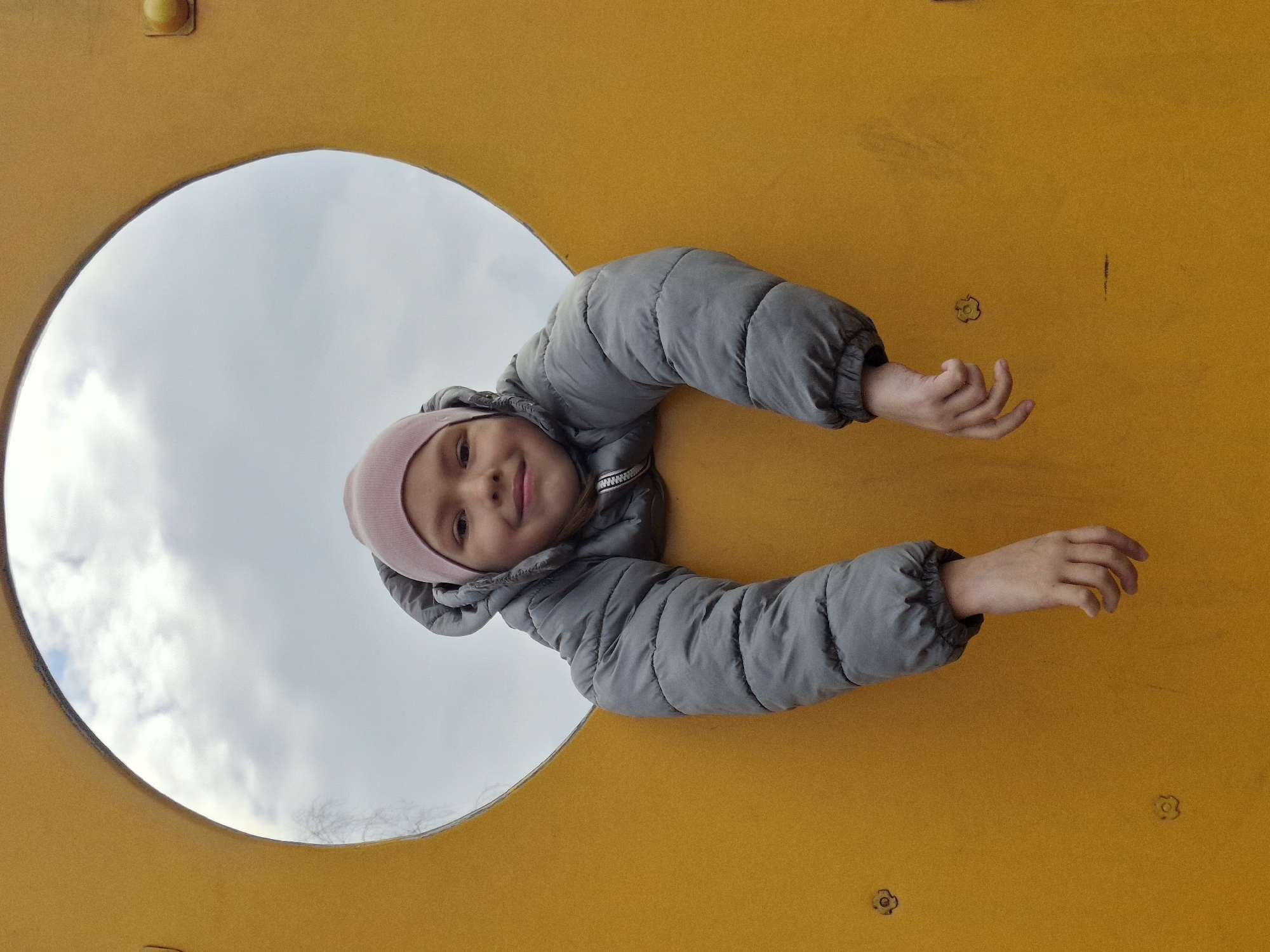 A young child looks directly at the camera through a circular opening in a bright yellow panel. The image is oriented with the child appearing horizontal, extending from the upper left to the right side of the frame, suggesting a 90-degree counter-clockwise rotation from an upright perspective. The child wears a grey padded winter jacket and a pink knitted beanie, and has a slight, pleasant smile. Both of the child's hands are visible, reaching out through the opening. Beyond the opening, a cloudy daytime sky is visible. The yellow panel, possibly part of a playground structure, shows some minor marks and wear. No text is visible in the image. The scene takes place in Chaikovsky, Russia.