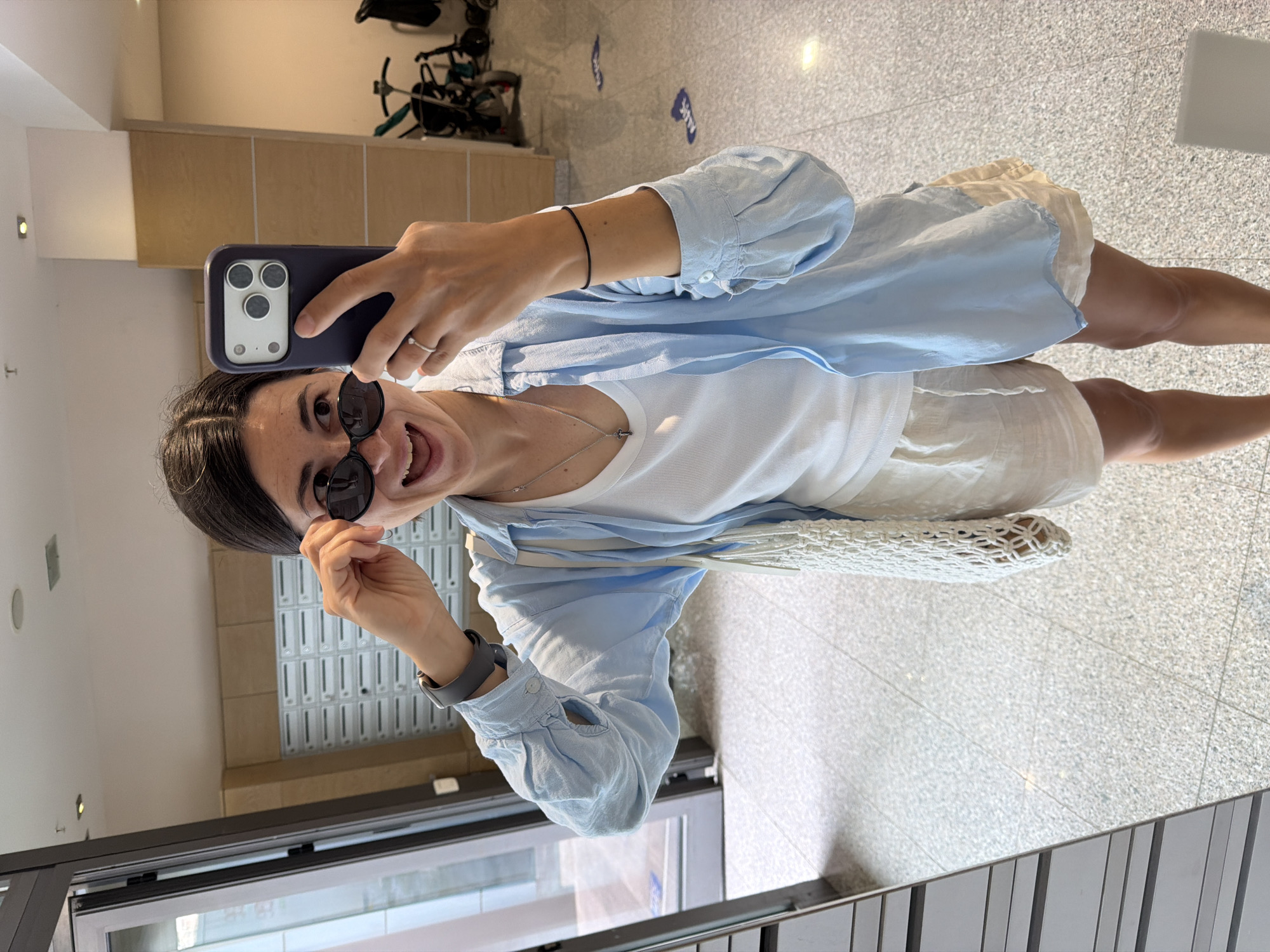 A woman stands indoors, posing for a selfie with her smartphone. She is wearing sunglasses and a blue shirt over a white t-shirt, along with shorts. A knit bag hangs from her shoulder. The location appears to be a building's lobby or entrance area, potentially in Budva, Montenegro, though this cannot be confirmed from visual evidence. The background includes mailboxes and a partial view of the exterior, possibly a street, and a bicycle parked inside. The floor is light-colored tile.