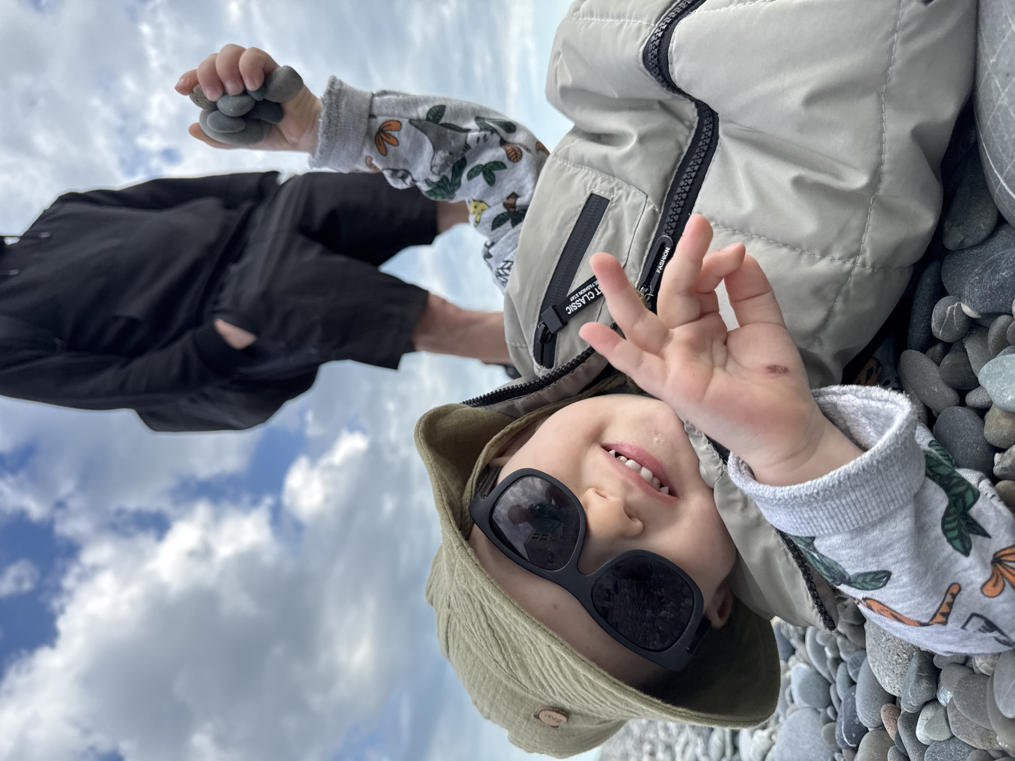 A child wearing sunglasses and a hat is lying on a pebble beach and smiling at the camera. The child is also wearing a grey jacket and a shirt with animal patterns. An adult wearing black clothing and holding stones is visible in the background. The background includes a cloudy sky. The location is likely a pebble beach in the city of Sochi, Russia.