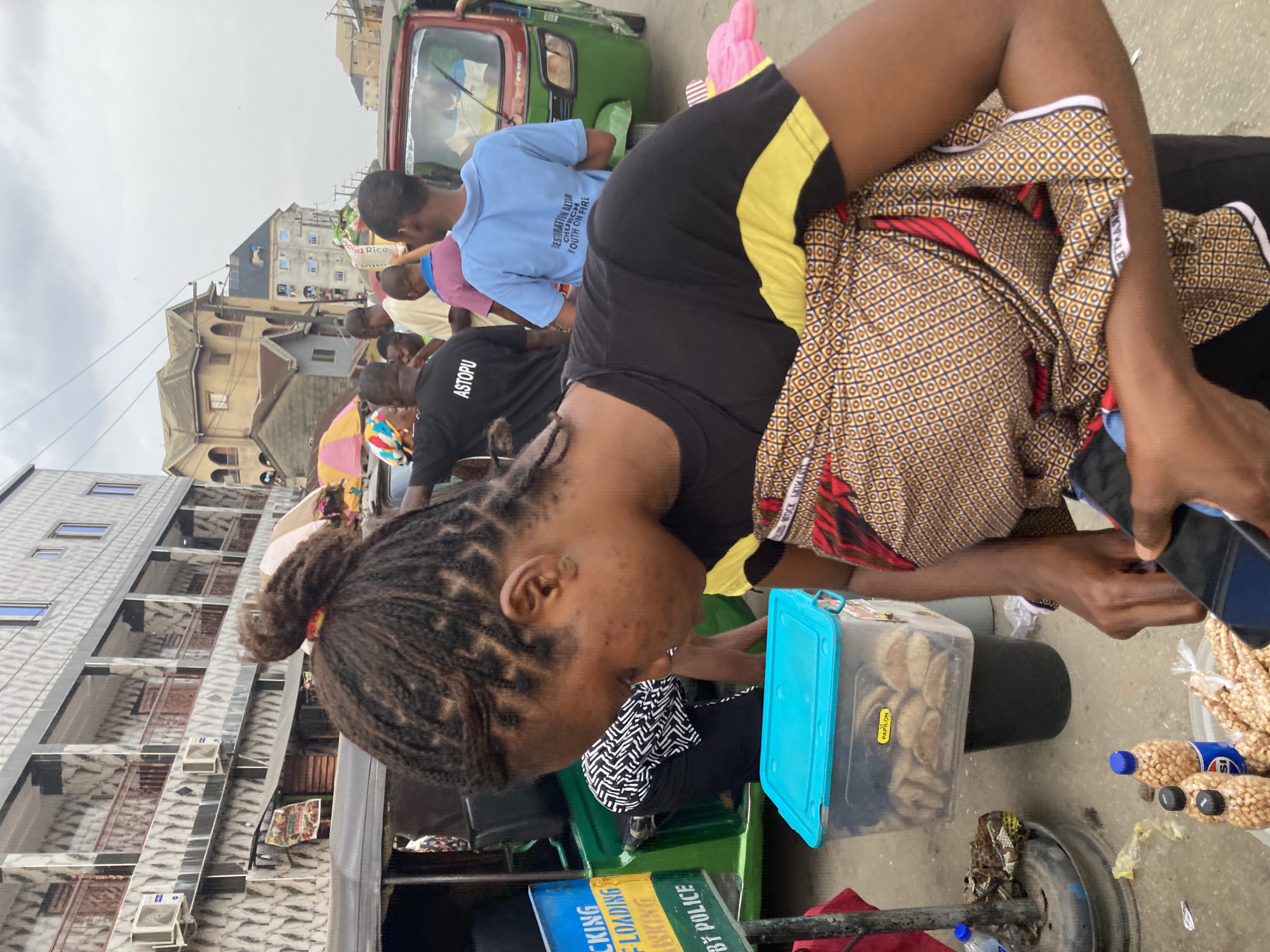 The image shows a woman with braided hair in Ura Nta Umuarandu, Nigeria. She is wearing a black and yellow shirt and patterned fabric. She stands near a blue container filled with what appears to be food. She holds a phone in her right hand. Nearby, there are bottles filled with peanuts. In the background, there is a green auto-rickshaw (Keke Napep), people walking, and buildings typical of an urban environment. A sign on the auto-rickshaw mentions "Debt Police" and "Loading."