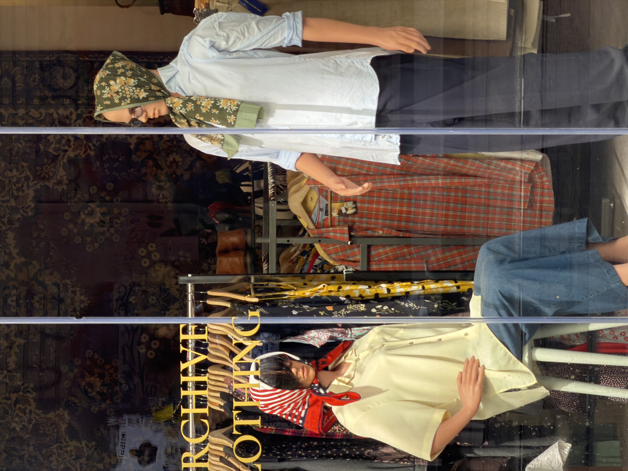 This image captures a window display of a clothing store, likely located in Sofia, Bulgaria, as suggested by the provided context. The scene is static, featuring two mannequins showcasing outfits through a glass pane, which also reflects internal elements of the shop.
One mannequin stands on the left, dressed in a light blue collared shirt with rolled-up sleeves, dark trousers, and a green headscarf adorned with yellow floral patterns. This mannequin also wears glasses and has its arms positioned as if in mid-gesture.
On the right, a second mannequin is seated on a simple white chair. It wears a light yellow short-sleeved button-up shirt, a blue denim skirt or wide-leg pants, and a distinctive red and white striped headscarf tied under its chin. Its hands rest casually on its lap.
Behind the mannequins, clothing racks are visible, laden with various garments, including a prominent red and black plaid shirt and other patterned fabrics. Wooden hangers can be seen. The background features a dark, intricately patterned curtain or wallpaper, possibly floral.
A notable detail is the reflection on the glass, which clearly displays the words "ARCHIVE CLOTHING" in vertically oriented, reflective gold letters, indicating the store's name or theme. The lighting suggests it is daytime, or the display is well-lit for viewing. No specific weather conditions or emotional expressions are discernible as the subjects are mannequins.
