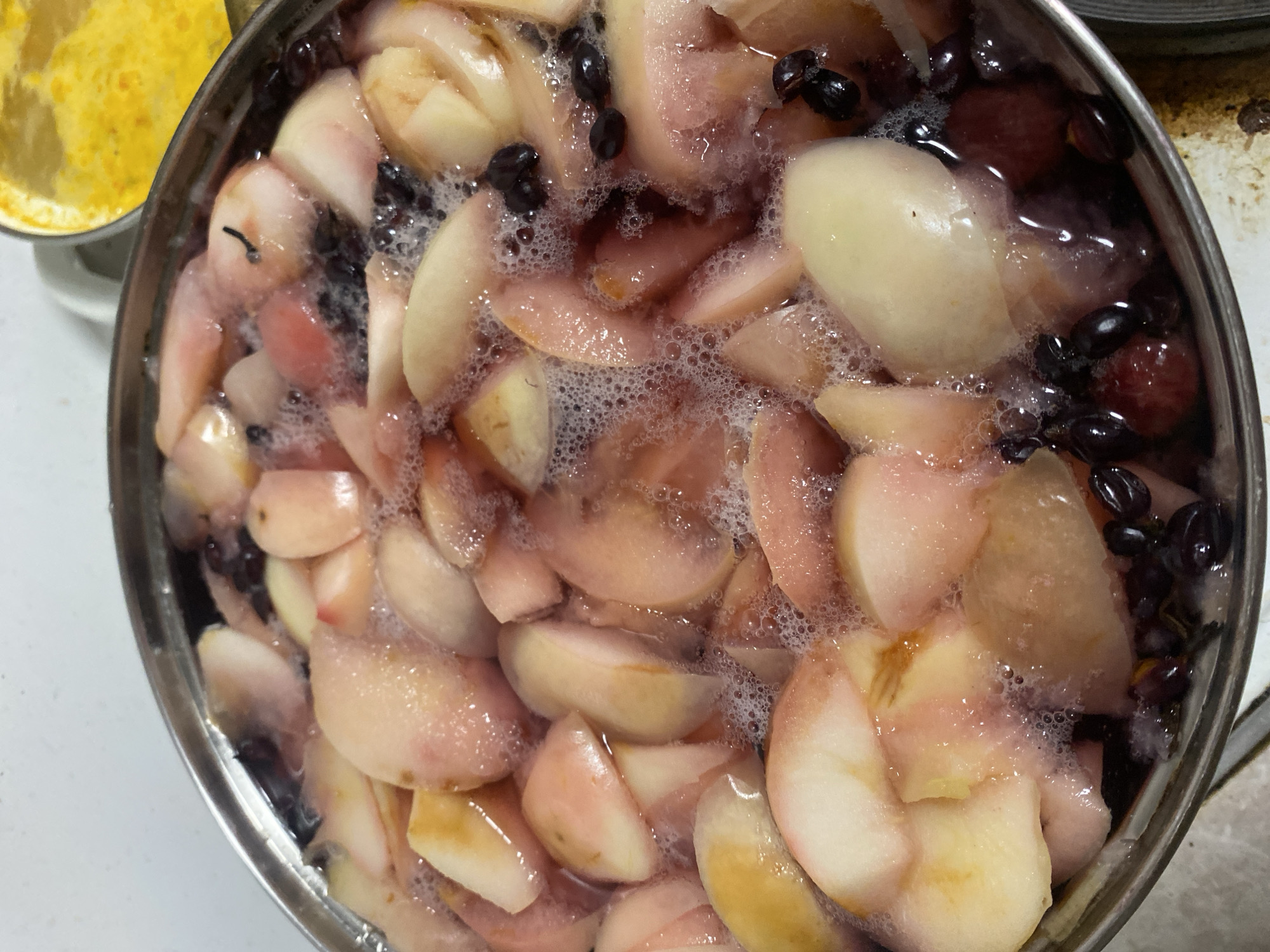An overhead close-up shot captures a pot of chopped peaches and dark berries, possibly blueberries or grapes, simmering in a liquid. The fruit is cut into irregular pieces and is submerged in a light pink, foamy liquid. Bubbles are visible on the surface, indicating the mixture is cooking. The pot appears to be made of stainless steel, with a visible rim. In the background, to the right, a yellow substance is visible in another container. The scene is likely in a kitchen or food preparation area in Chornomorsk, Ukraine.