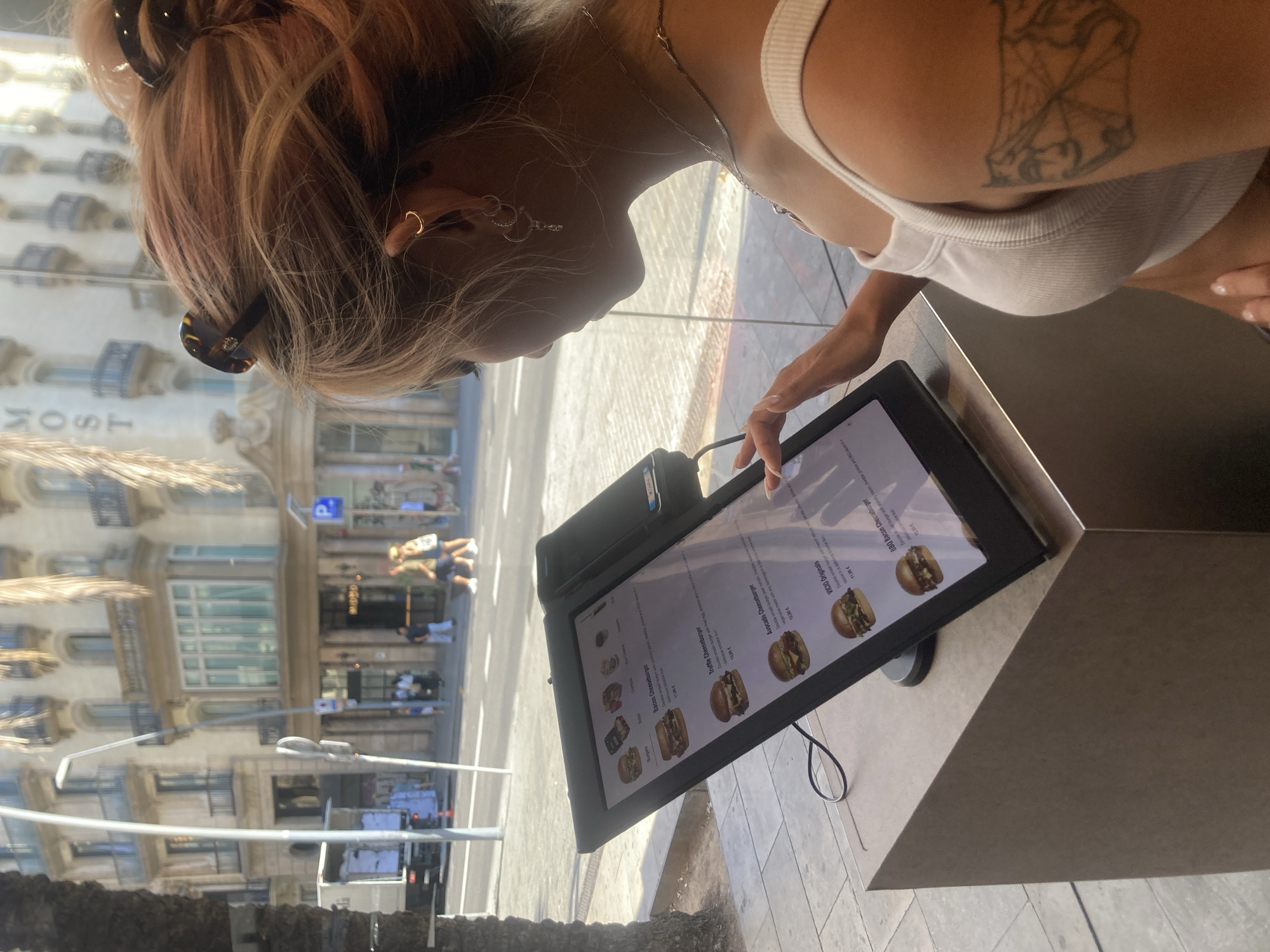 A young woman with pink-tinted hair pulled back in a bun is seen from the side, interacting with a touchscreen menu display. The display shows a menu for a burger restaurant, with images of various burgers and their descriptions, including "Bacon Cheeseburger," "Truffle Cheeseburger," and "Avocado Cheeseburger." The woman is wearing a white tank top and has tattoos on her arm. She is standing at an outdoor counter, likely in a cafe or restaurant, with a view of a city street and buildings through a large window. The background suggests a European city, with architectural styles consistent with Barcelona, Spain, including ornate building facades and balconies. The street appears busy with pedestrians and a delivery truck. The overall scene captures a moment of casual dining or ordering.