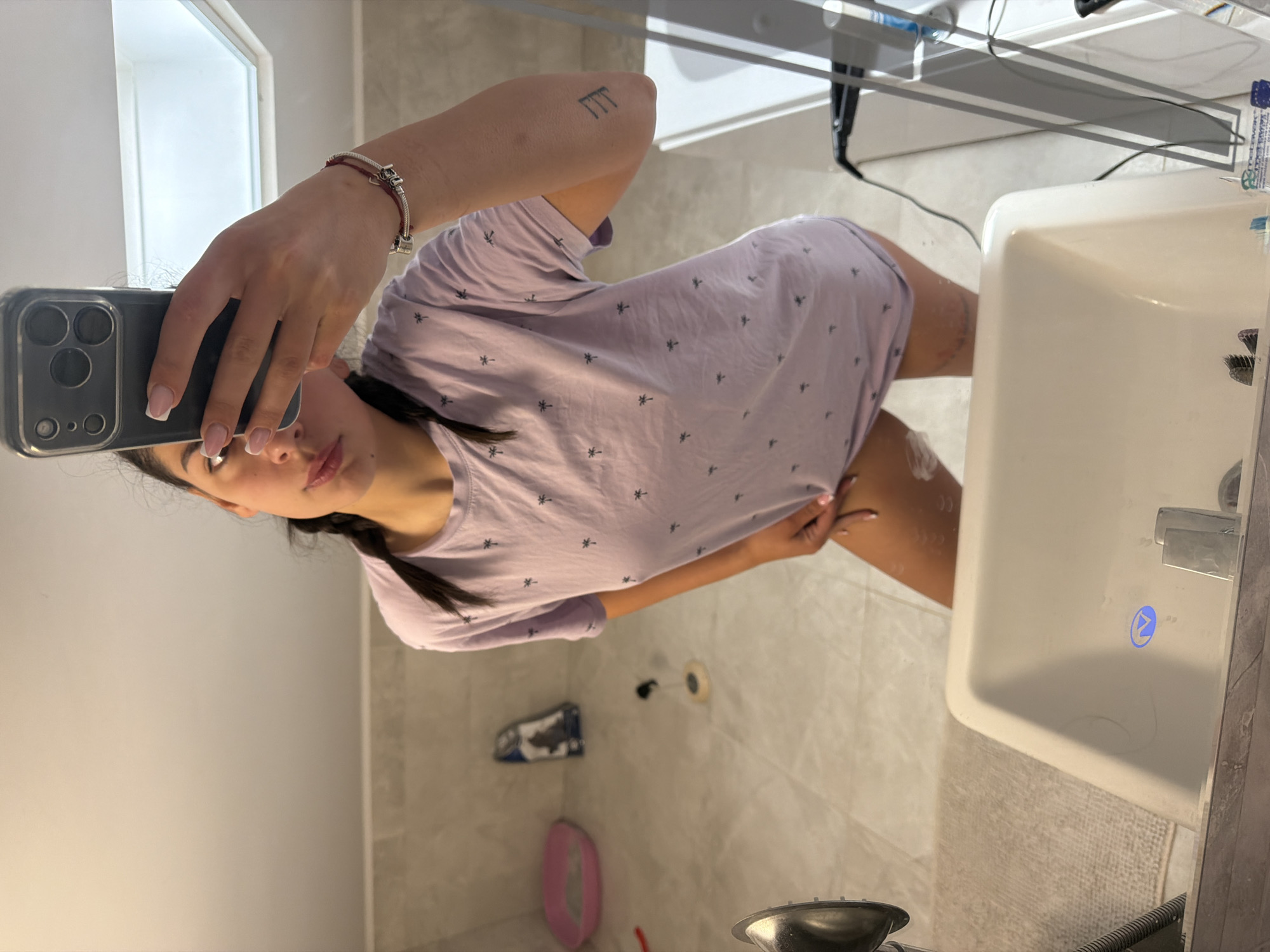 The image shows a person in a bathroom taking a selfie. The person has brown hair in a braid and is wearing a purple t-shirt with palm tree designs. The phone being used to take the selfie is an iPhone, with its camera clearly visible. A silver bracelet is on the person's left wrist. The bathroom is tiled, with a white sink visible on the right side of the frame. A hair dryer is seen near the sink. Other bathroom items are scattered around the scene. The location is unknown, and nothing indicates Gabrovo, Bulgaria.